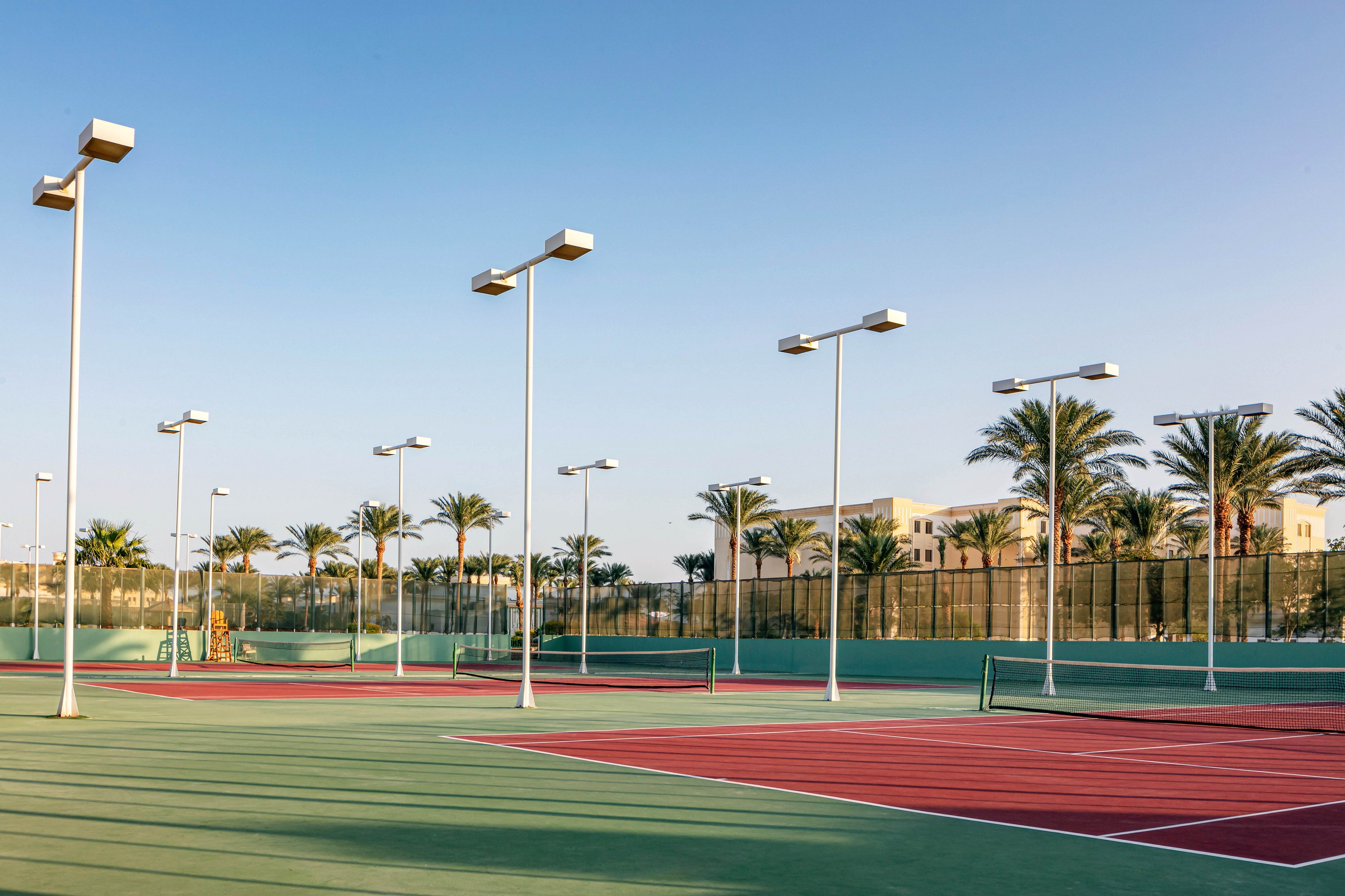 The image features several outdoor tennis courts surrounded by tall palm trees and modern lighting poles. The courts are empty, with red and green surfaces, and the setting appears to be a warm, sunny climate. Buildings and fencing are visible in the background, and the sky is clear with a soft evening light. No visible text or numeric values are present.