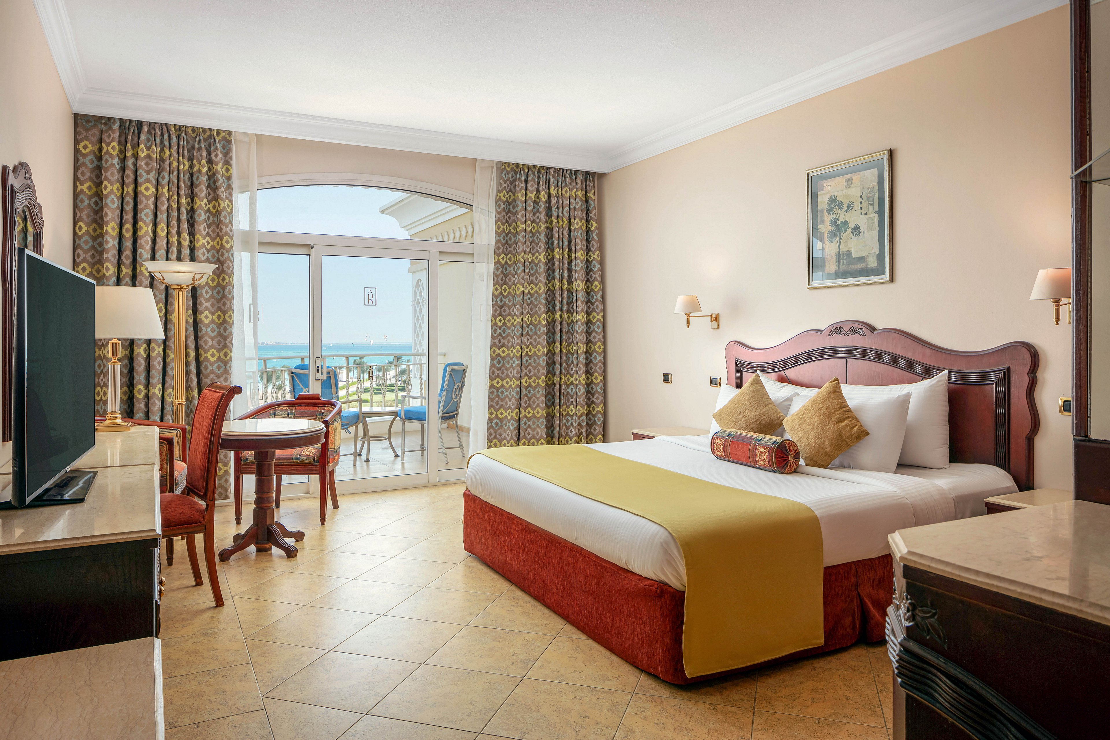 A spacious hotel room features a large bed with decorative pillows and a yellow throw, a wooden table with chairs, and a flat-screen TV. The room opens to a balcony overlooking the sea, with patterned curtains framing the glass doors. Warm neutral tones and classic furnishings create a welcoming and comfortable atmosphere.