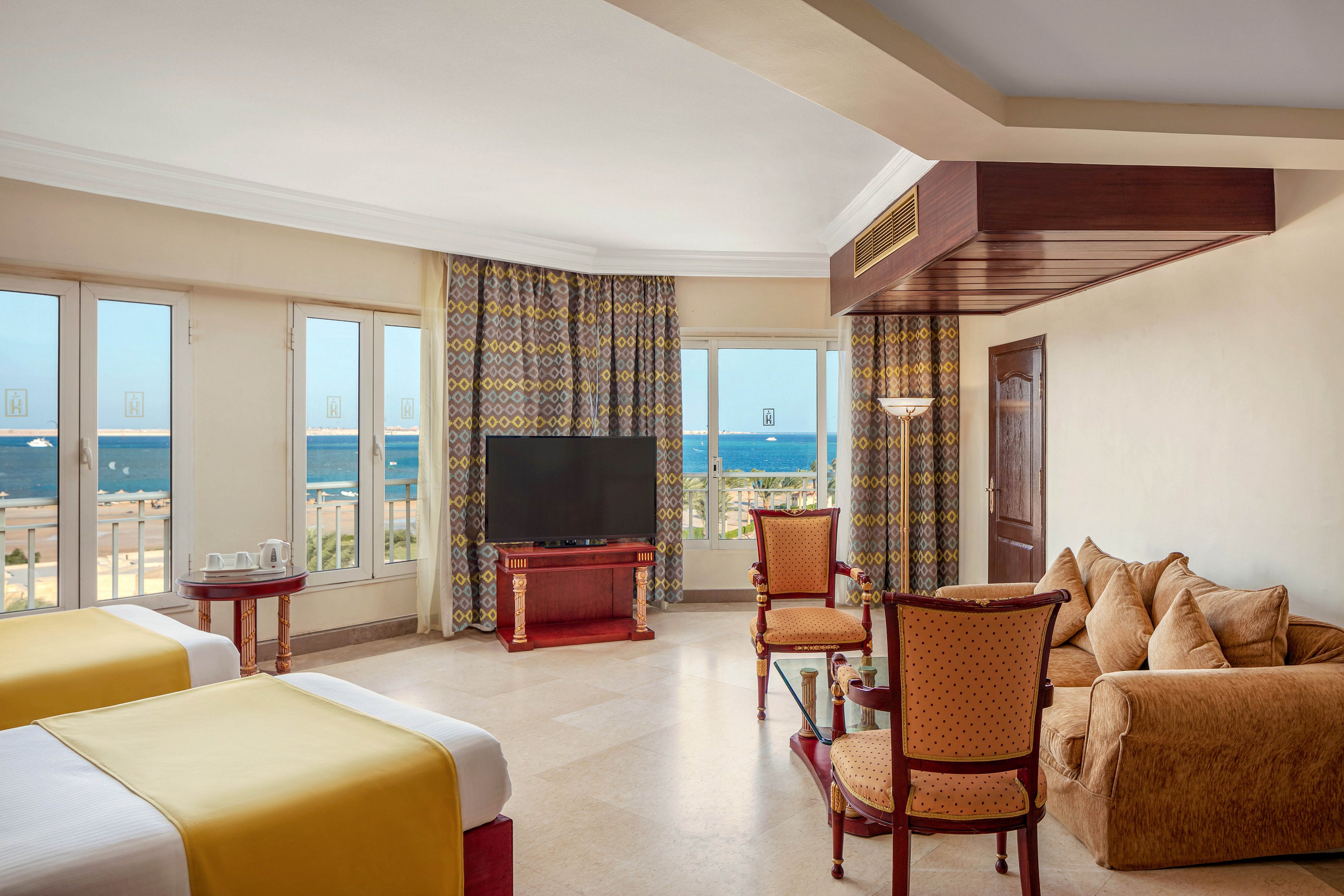 A large hotel room features two twin beds with yellow bed runners, a seating area with chairs and a sofa, and a flat-screen television. Multiple windows offer a clear view of the ocean, filling the space with natural light. The room is decorated in warm tones with patterned curtains and light-colored walls, creating a welcoming and relaxed atmosphere.