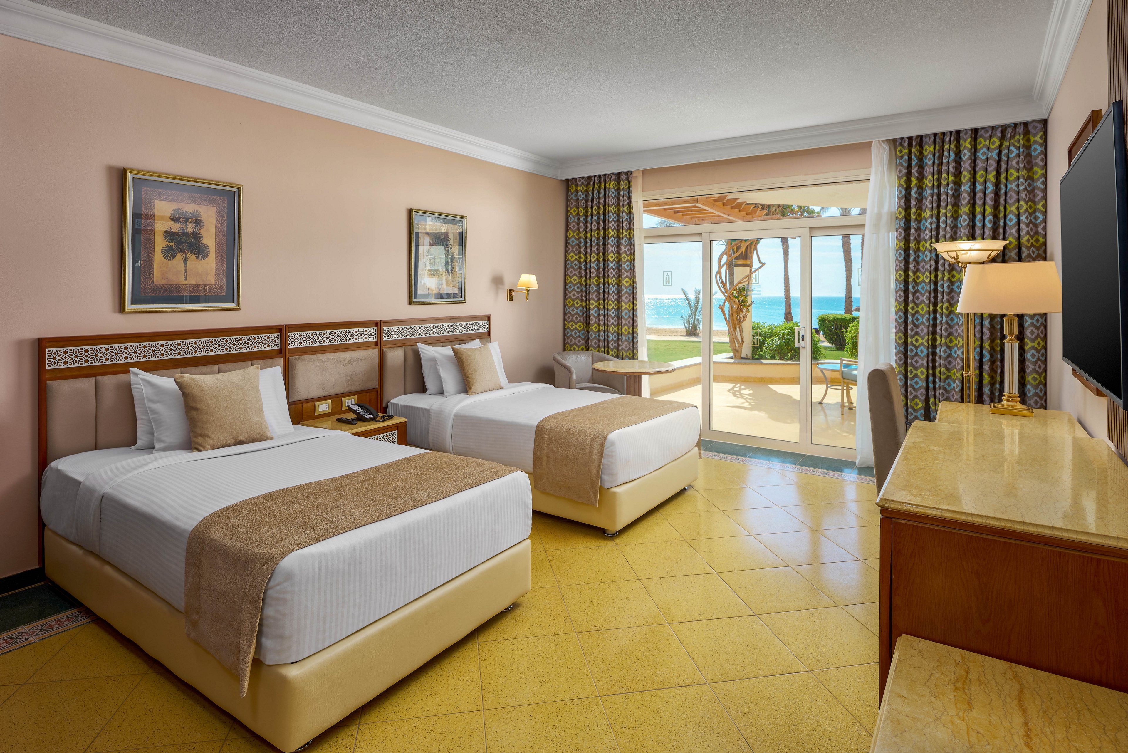 The image shows a spacious hotel room featuring two neatly made twin beds with beige and white bedding. The room has a large window and glass door leading to a patio with a view of the ocean and palm trees. Decor includes framed artwork, patterned curtains, and a desk with a television. The overall mood is bright and inviting, with a warm yellow tile floor and natural light.