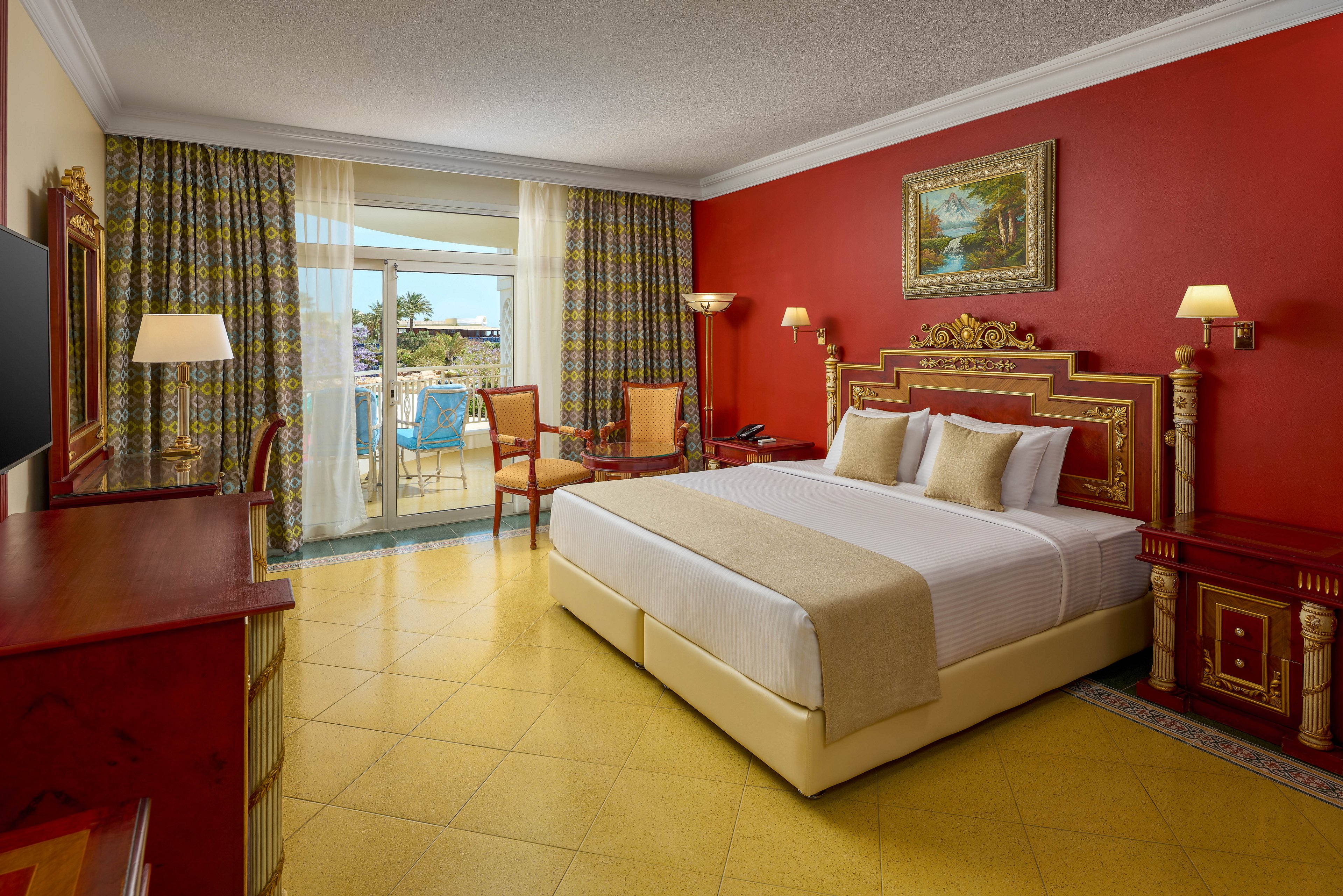 A spacious hotel room features a large king-size bed with gold-accented headboard and neutral bedding. The room is decorated in warm red and yellow tones, with classic wooden furniture and a painting above the bed. Large windows with patterned curtains open to a balcony overlooking an outdoor area, allowing natural light to fill the space. The setting is inviting and comfortable, ideal for relaxation or vacation stays.