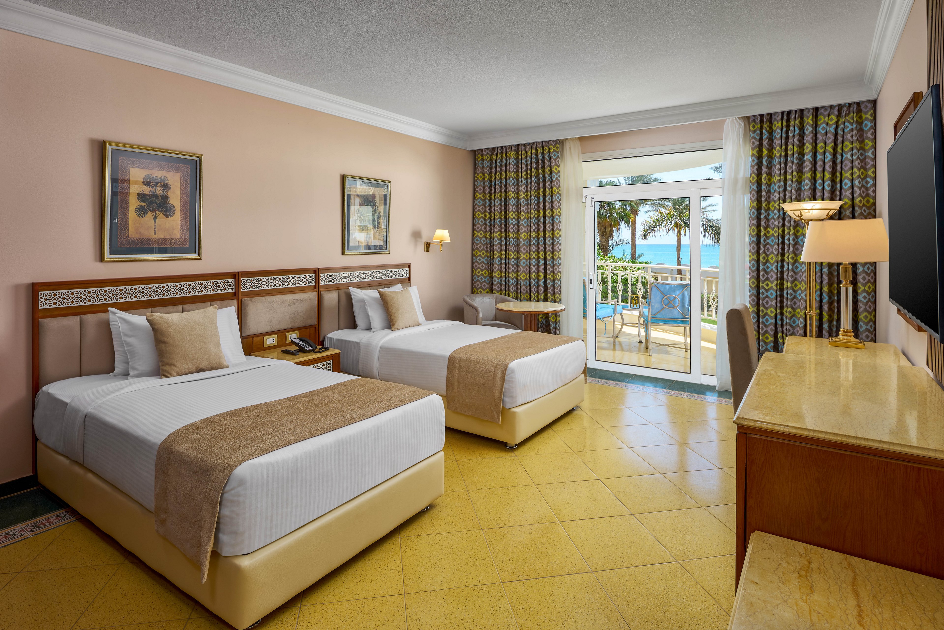A spacious hotel room features two neatly made twin beds with beige and white bedding. The room is decorated with framed artwork, patterned curtains, and a large desk facing a flat-screen TV. Sunlight streams in through glass doors leading to a balcony with outdoor seating and a view of palm trees and the ocean. The overall atmosphere is clean, inviting, and relaxed.