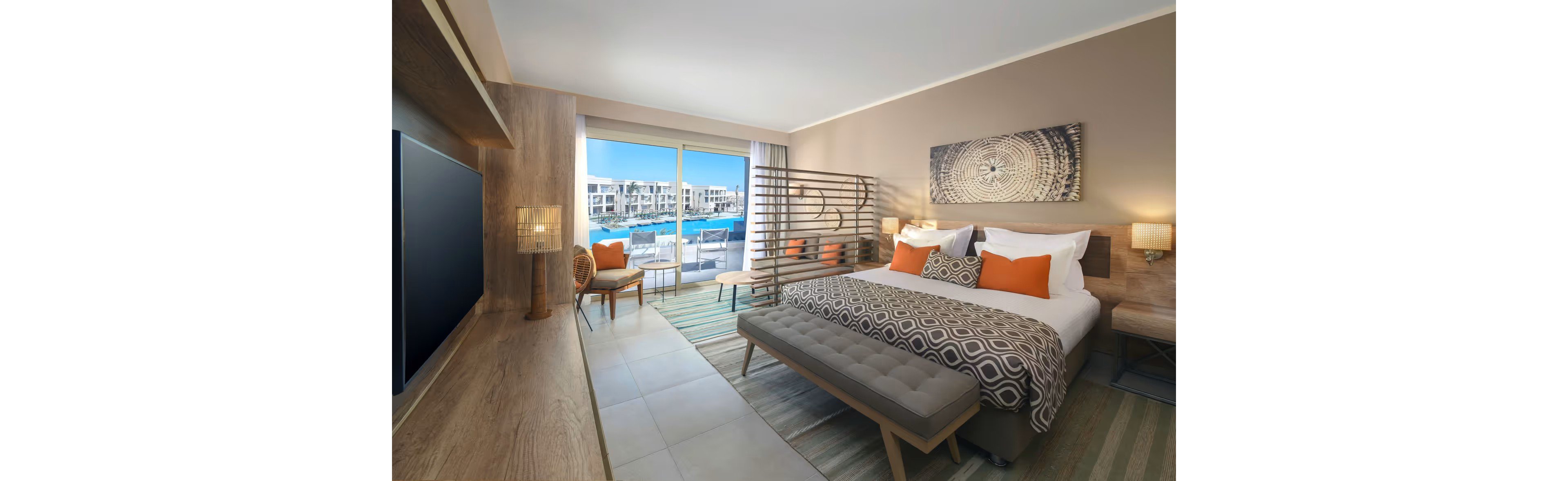 Contemporary hotel room features a large bed with patterned bedding and orange accent pillows. A cushioned bench sits at the foot of the bed, with a decorative partition separating the sleeping area from a small seating space. Floor-to-ceiling sliding glass doors open to a balcony with chairs and a view of an expansive resort pool and surrounding buildings. Neutral tones, wood finishes, and soft lighting create a calm, upscale atmosphere.
