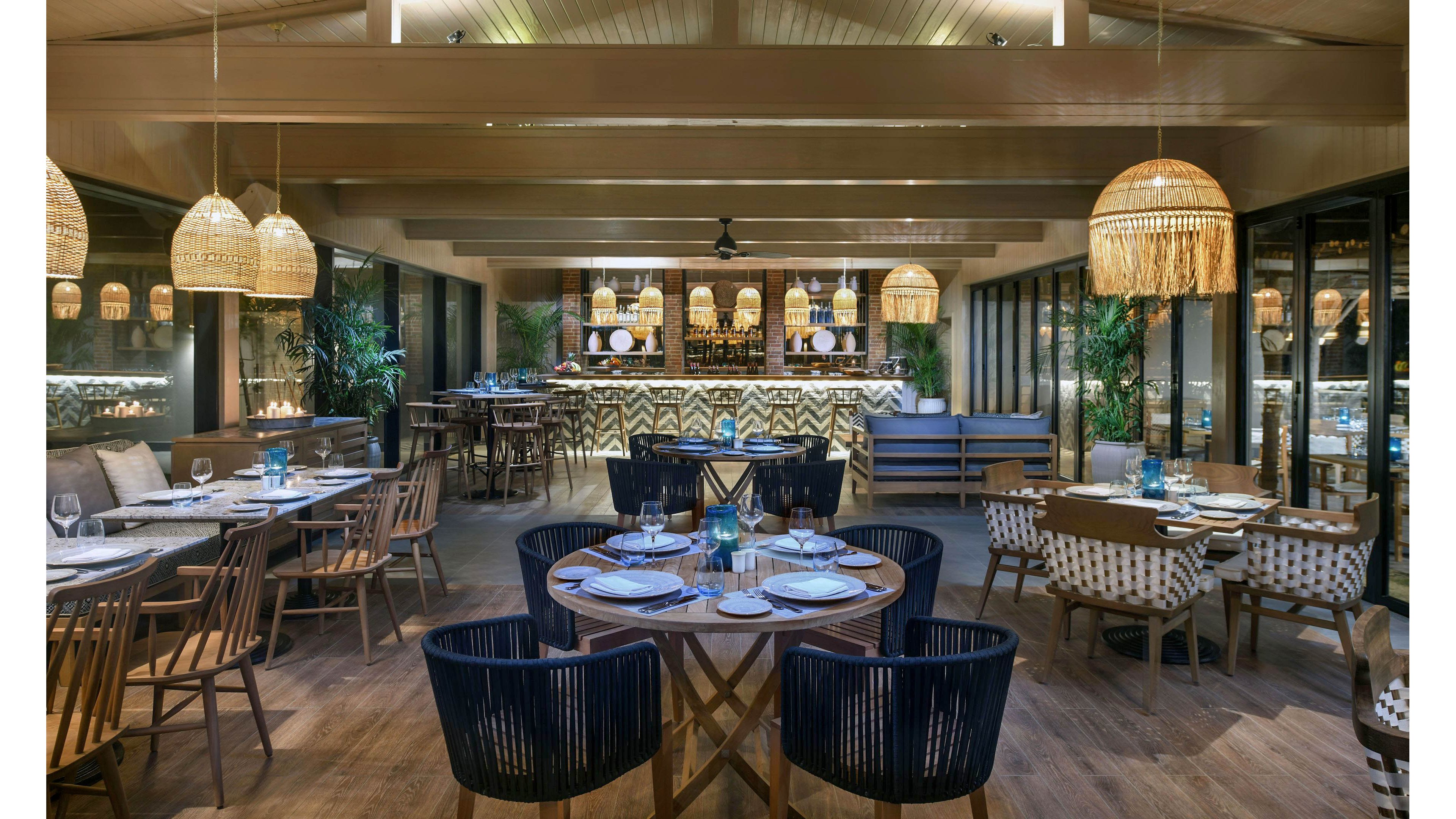 A stylish restaurant dining area features neatly set tables and a well-lit bar in the background. The space is decorated with woven pendant lights, wooden furniture, and lush green plants, creating a warm and inviting atmosphere. The overall palette is neutral with natural wood tones and soft lighting, enhancing the cozy and contemporary vibe. No visible people or text are present in the image.