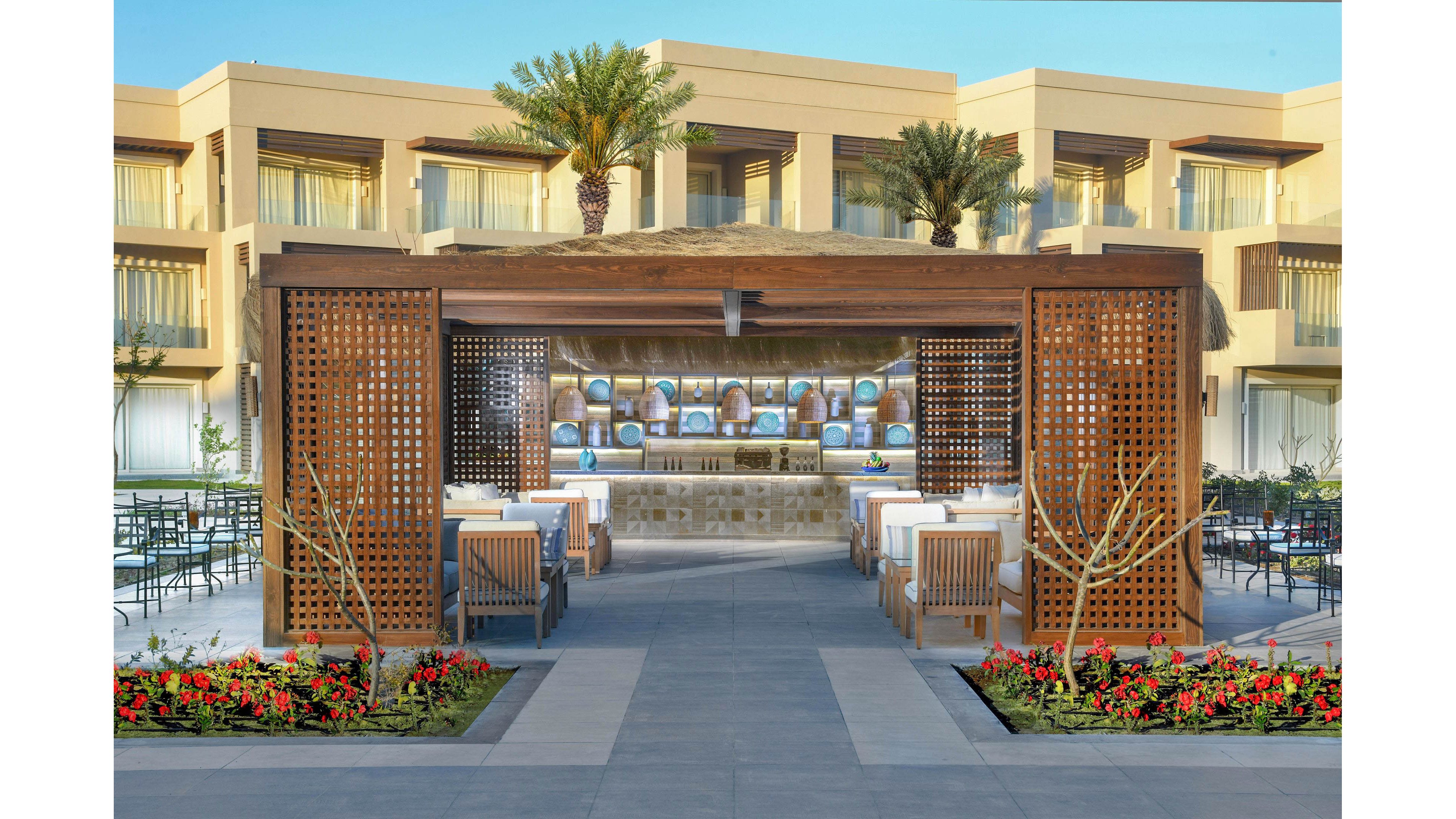 A contemporary outdoor bar is featured at the center of a resort courtyard, surrounded by modern seating and lush landscaping. The setting includes palm trees, vibrant flower beds, and a backdrop of beige hotel buildings with balconies. The bar area is shaded by a wooden pergola with decorative lattice panels, creating a relaxed and inviting atmosphere. The overall mood is bright and welcoming, ideal for leisure and social gatherings.