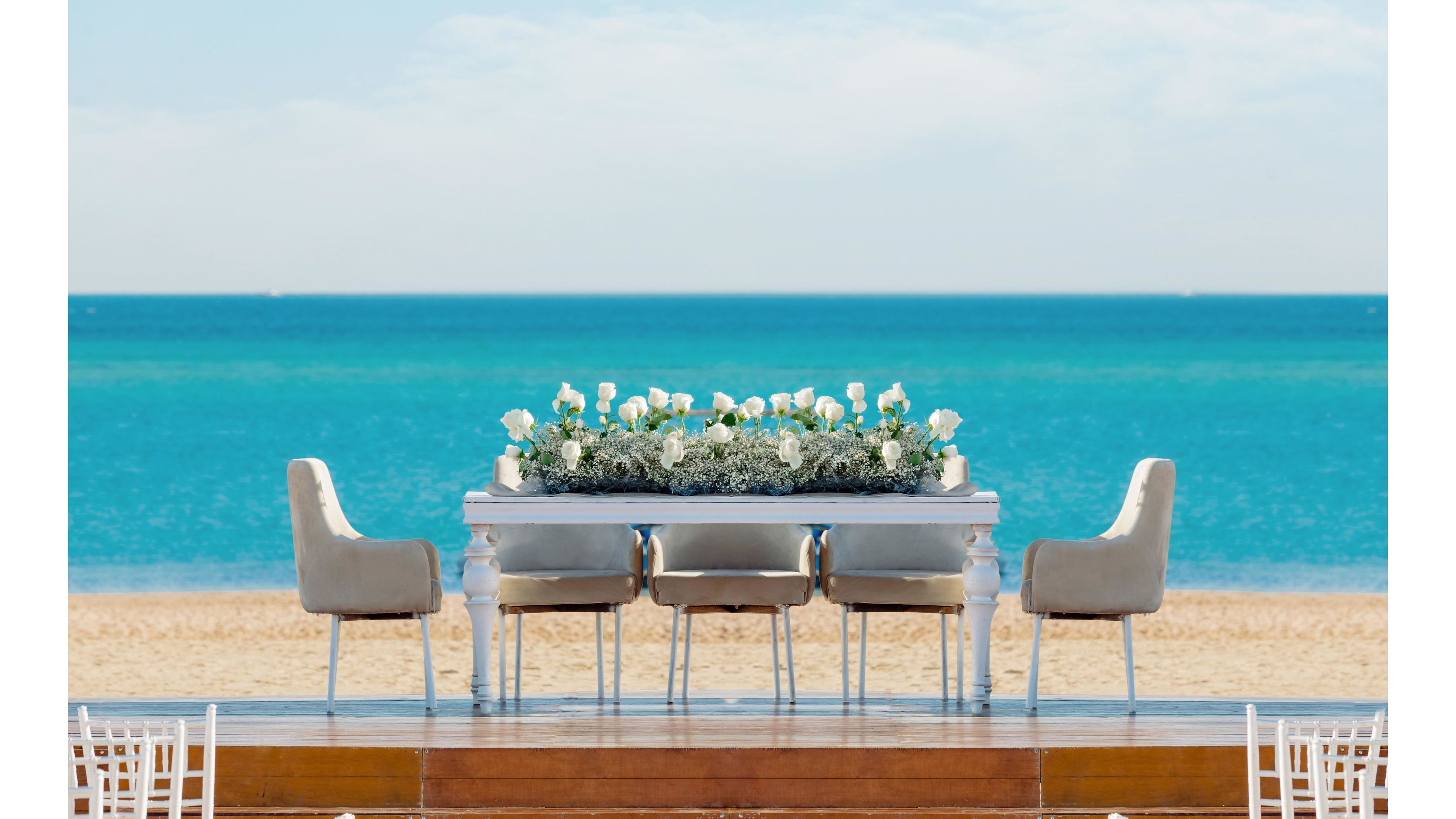 A sophisticated dining table with plush chairs is arranged on a wooden platform overlooking a serene blue ocean. The table features a lush white floral centerpiece, creating a luxurious and inviting atmosphere. The scene is set on a sandy beach under a clear sky, evoking a tranquil and celebratory mood. No visible text or numbers are present in the image.