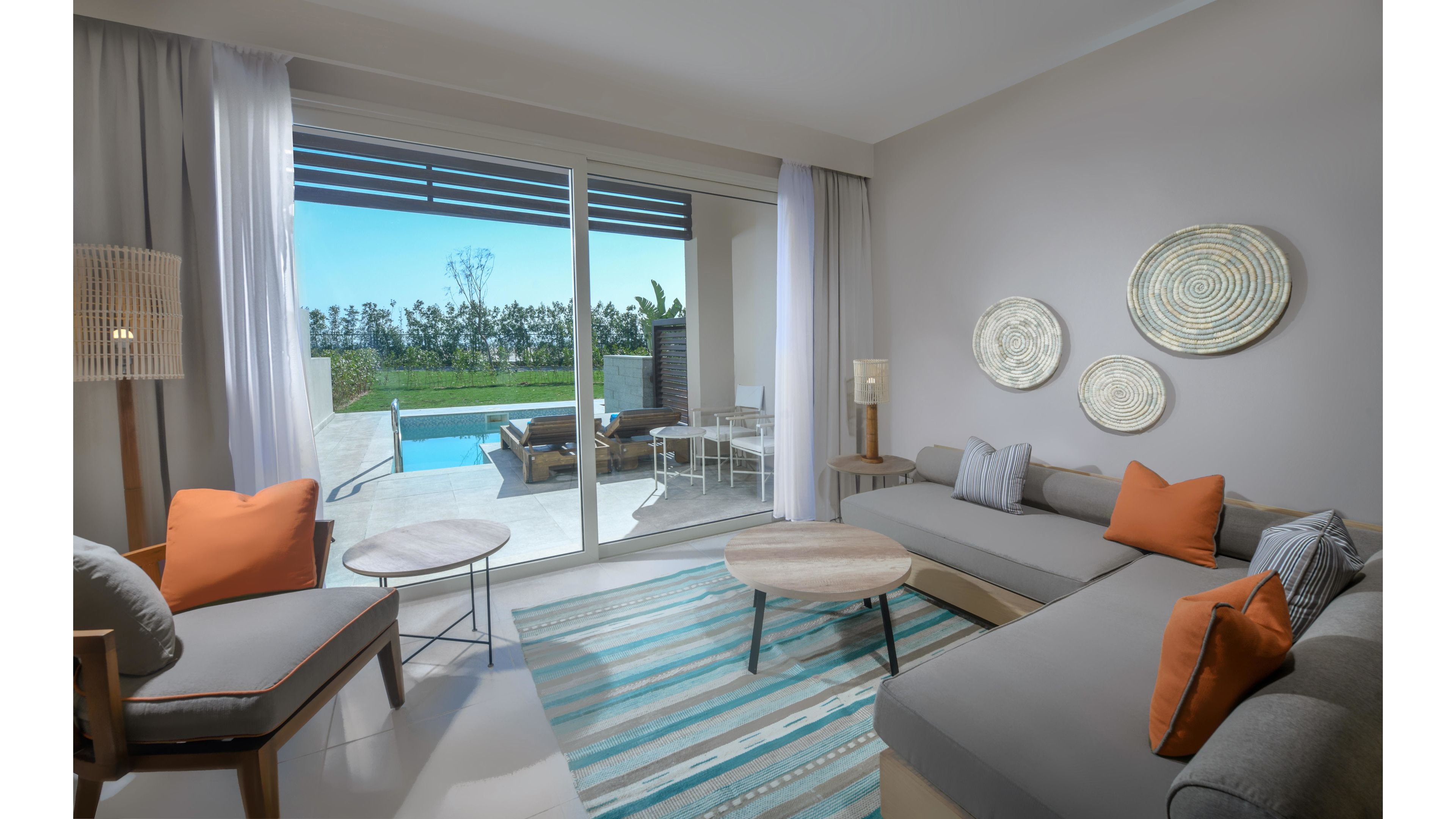 A contemporary resort suite features a spacious seating area with neutral tones and orange accent pillows. Large sliding glass doors open to a private patio and pool, offering a view of lush greenery. The room is decorated with woven wall art and a striped area rug, creating a relaxed and inviting atmosphere. Natural light fills the space, highlighting the clean and modern interior design.