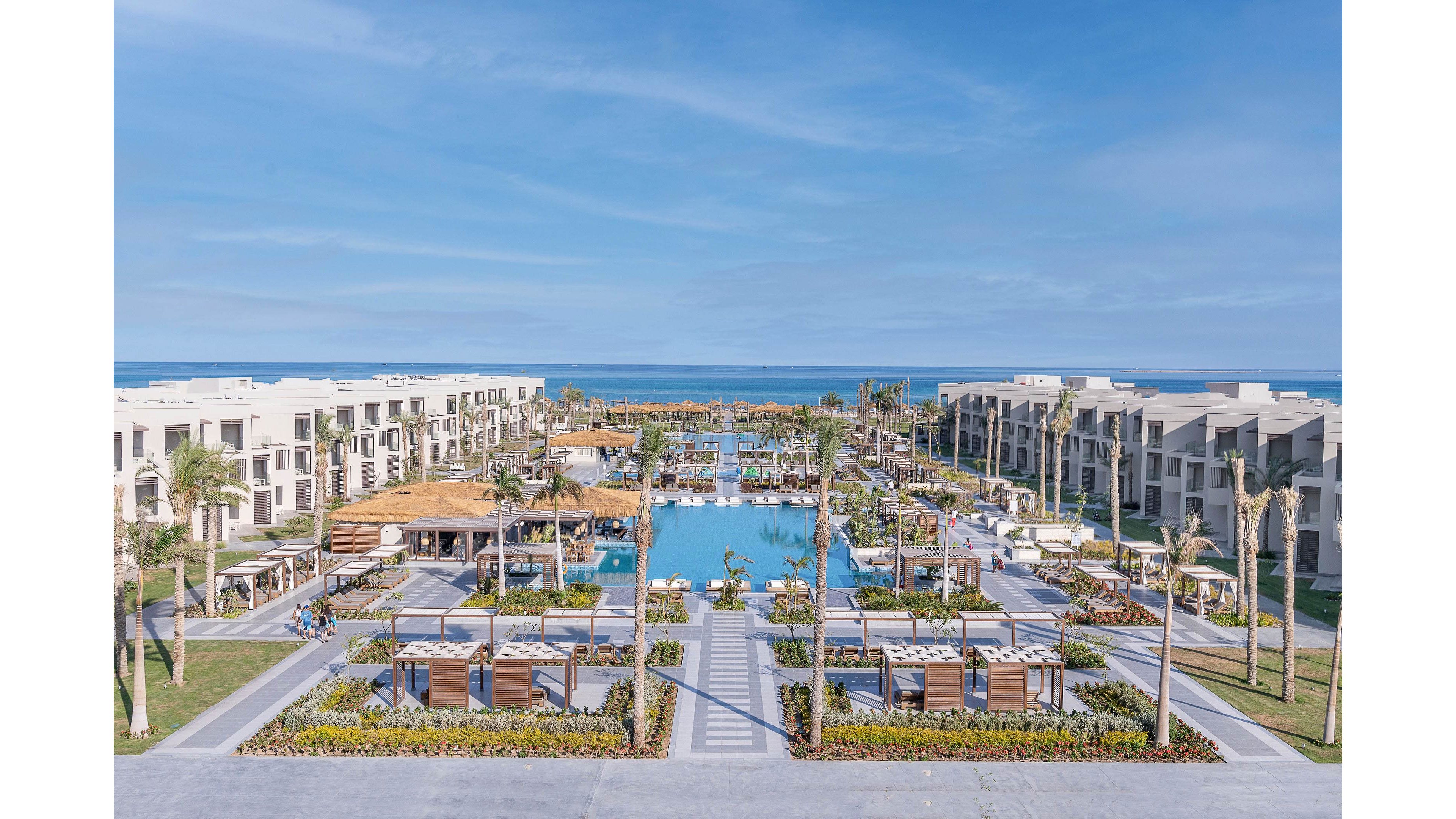 A modern beachfront resort features a large central swimming pool surrounded by palm trees, cabanas, and landscaped gardens. The setting is tranquil and inviting, with clear blue skies and the ocean visible in the background. The architecture is contemporary, with white and beige buildings lining both sides of the pool. The overall mood is relaxed and upscale, ideal for vacation or leisure.