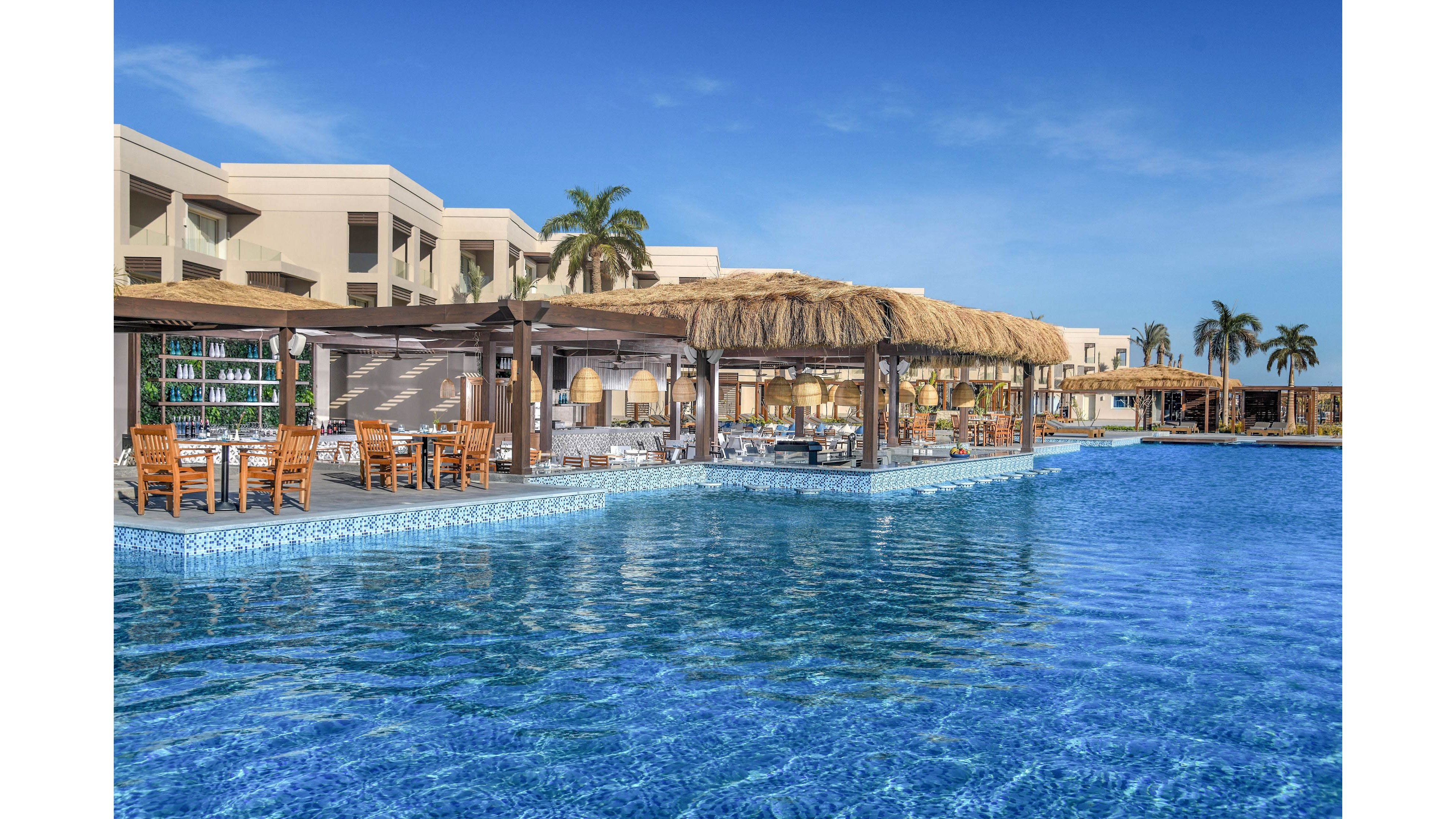 A spacious outdoor restaurant with wooden furniture and thatched roofs sits beside a large, inviting swimming pool. The setting features modern resort architecture, palm trees, and clear blue skies, evoking a relaxed vacation atmosphere. The pool's shimmering water and open-air dining area create a tropical, leisurely mood. No visible text or numbers are present in the image.