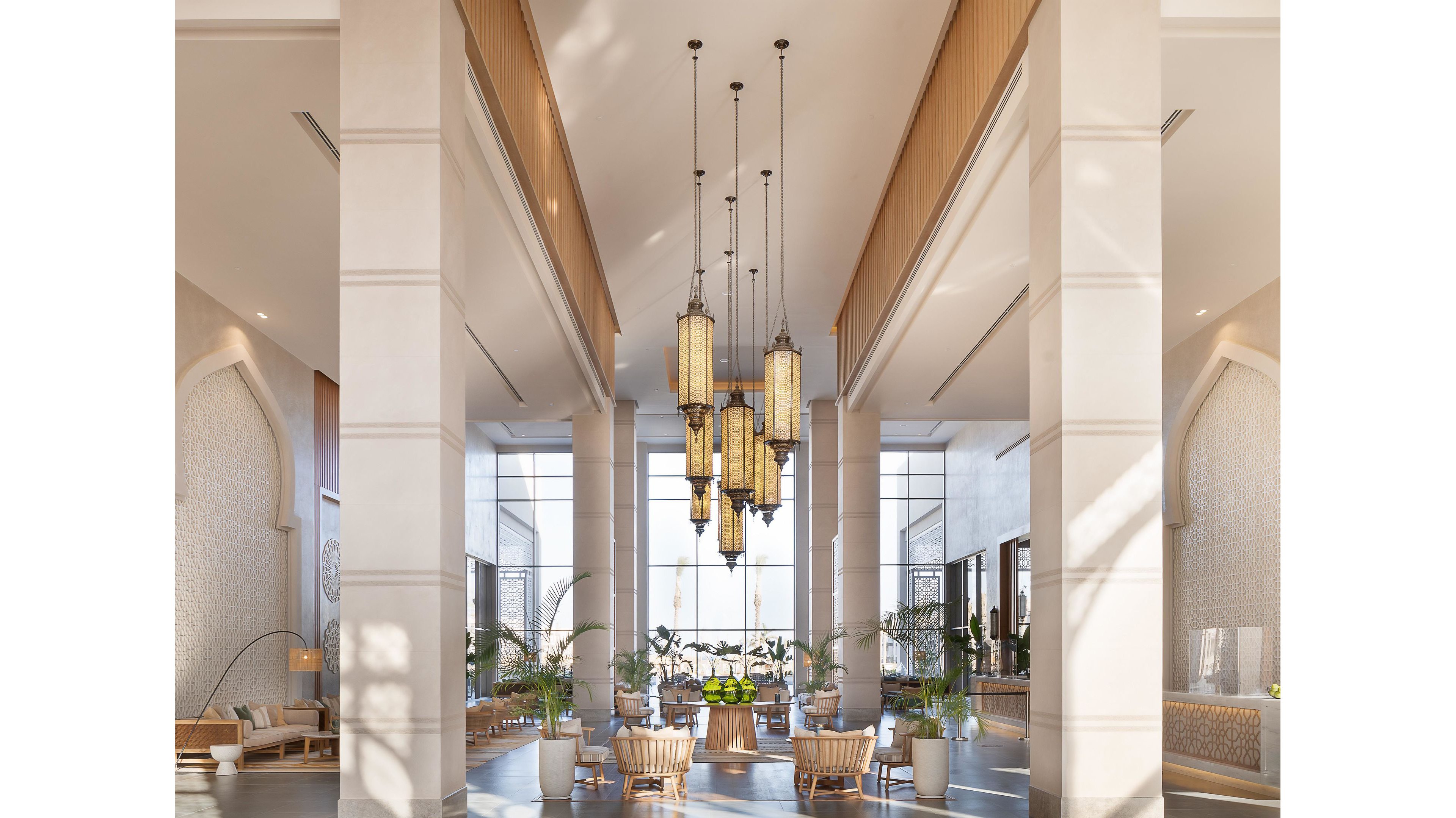 A spacious and luxurious hotel lobby features tall columns, large hanging chandeliers, and abundant natural light. The seating area is arranged with modern chairs and tables, surrounded by potted plants and decorative elements. The neutral color palette and high ceilings create an airy, welcoming atmosphere, with intricate wall details adding sophistication.