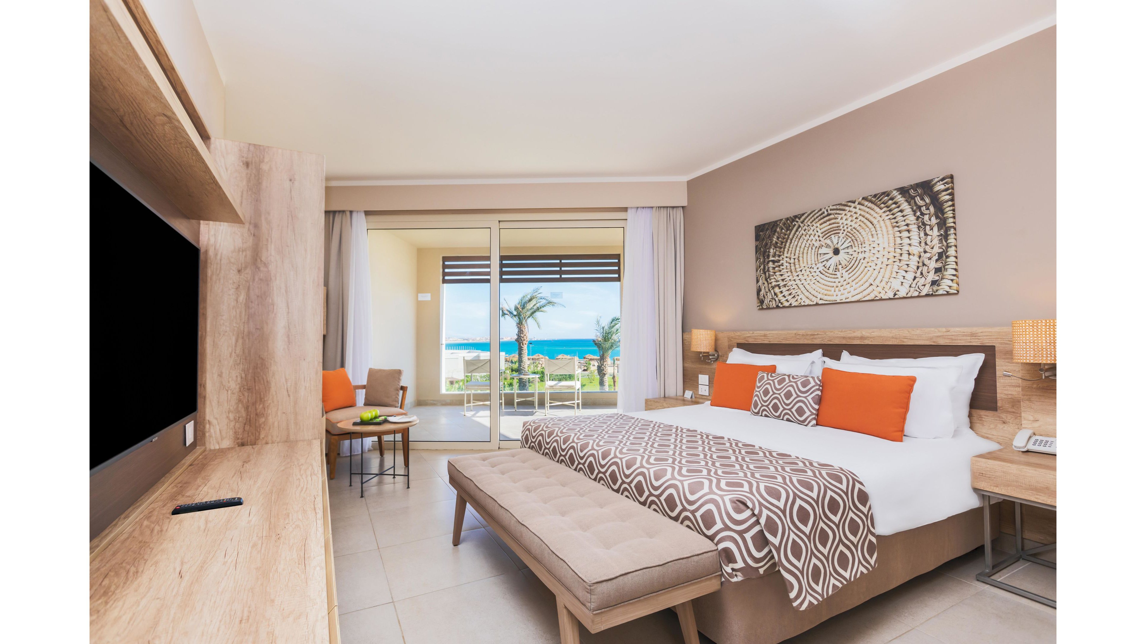 A contemporary hotel room features a large bed with geometric-patterned bedding and orange accent pillows. The room opens to a balcony overlooking palm trees and a blue ocean, creating a relaxing vacation atmosphere. Neutral tones and natural light enhance the inviting, modern style, while a wall-mounted TV and cozy seating area add comfort. No visible text or numbers are present in the image.