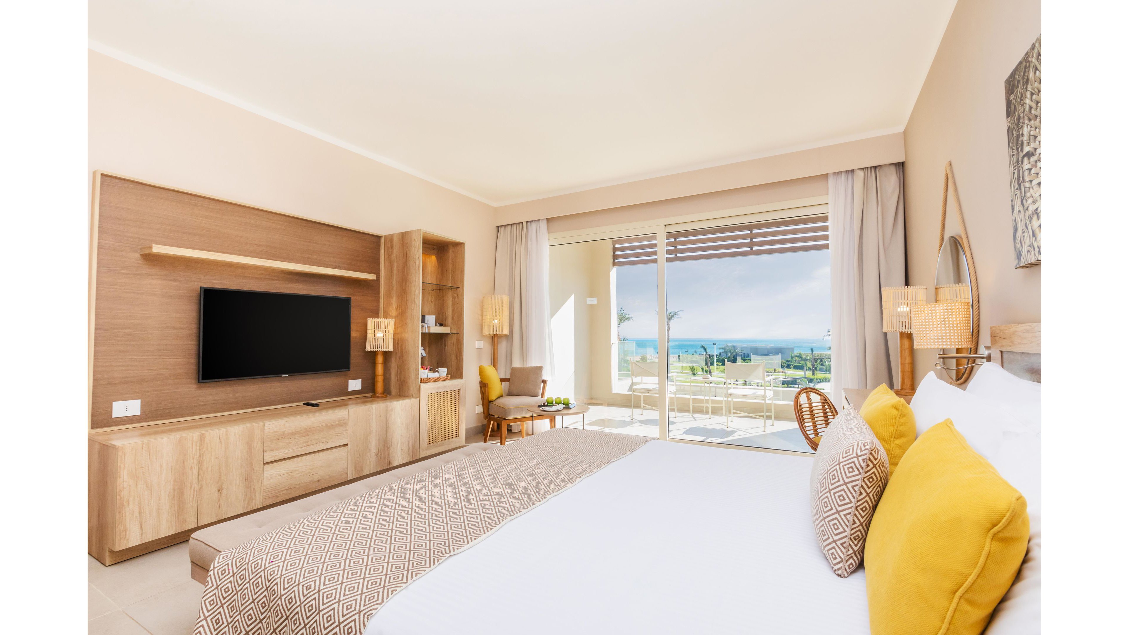 A spacious, contemporary hotel room features a large bed with patterned pillows and a wooden headboard. The room includes a flat-screen TV, built-in shelving, and a cozy seating area near a glass door leading to a balcony with a scenic ocean view. Natural light fills the space, highlighting the neutral color palette and minimalist decor. The setting is bright and inviting, ideal for relaxation or vacation stays.