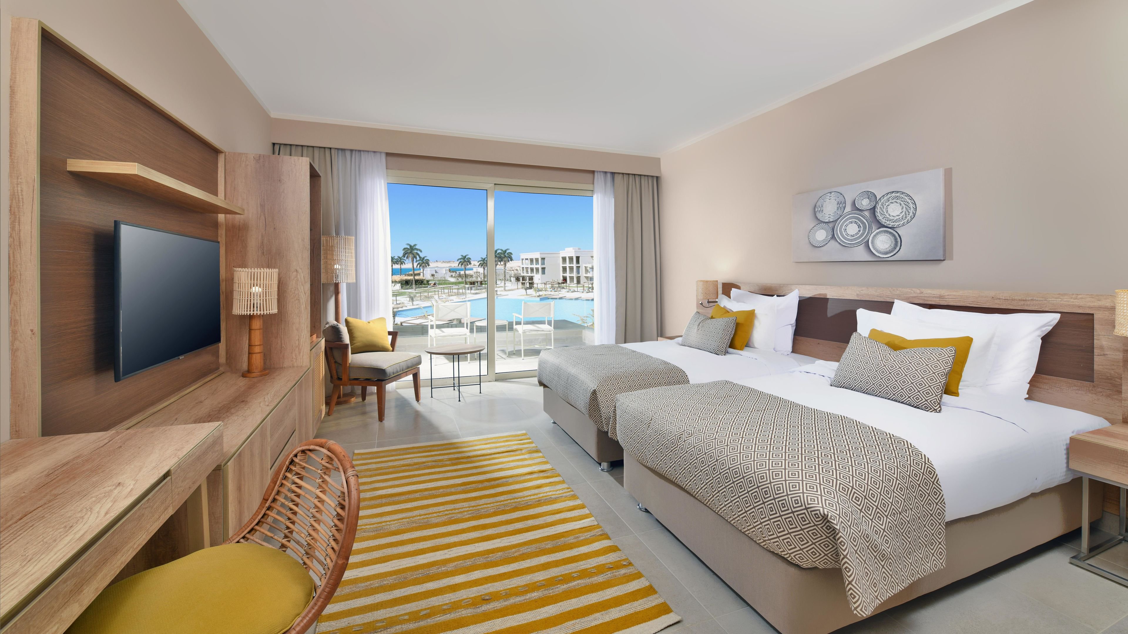A contemporary hotel room features two neatly made twin beds with yellow and gray accents. The space includes a wooden desk, flat-screen TV, and a cozy seating area by the window. Large glass doors open to a balcony overlooking a pool and resort area. The room is decorated in a neutral palette with pops of yellow, creating a bright and inviting atmosphere. No visible text or numbers are present in the image.