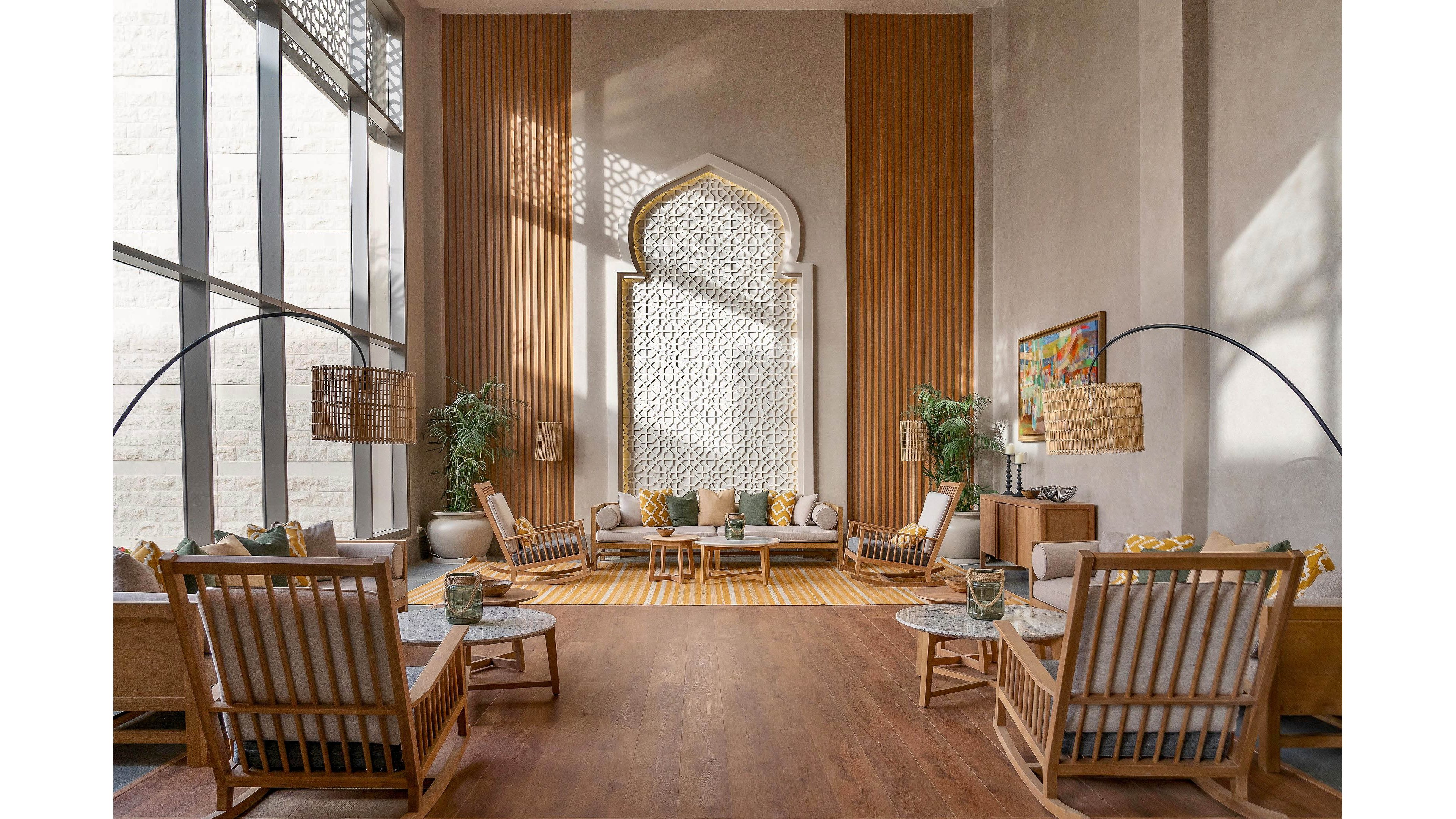 A spacious, sunlit lounge features contemporary wooden seating arranged around a central coffee table. The room is accented by a large, ornate arched wall detail and tall windows that let in natural light. Neutral tones and yellow accents create a warm, inviting atmosphere, complemented by potted plants and modern floor lamps. The setting is elegant and serene, ideal for relaxation or social gatherings.