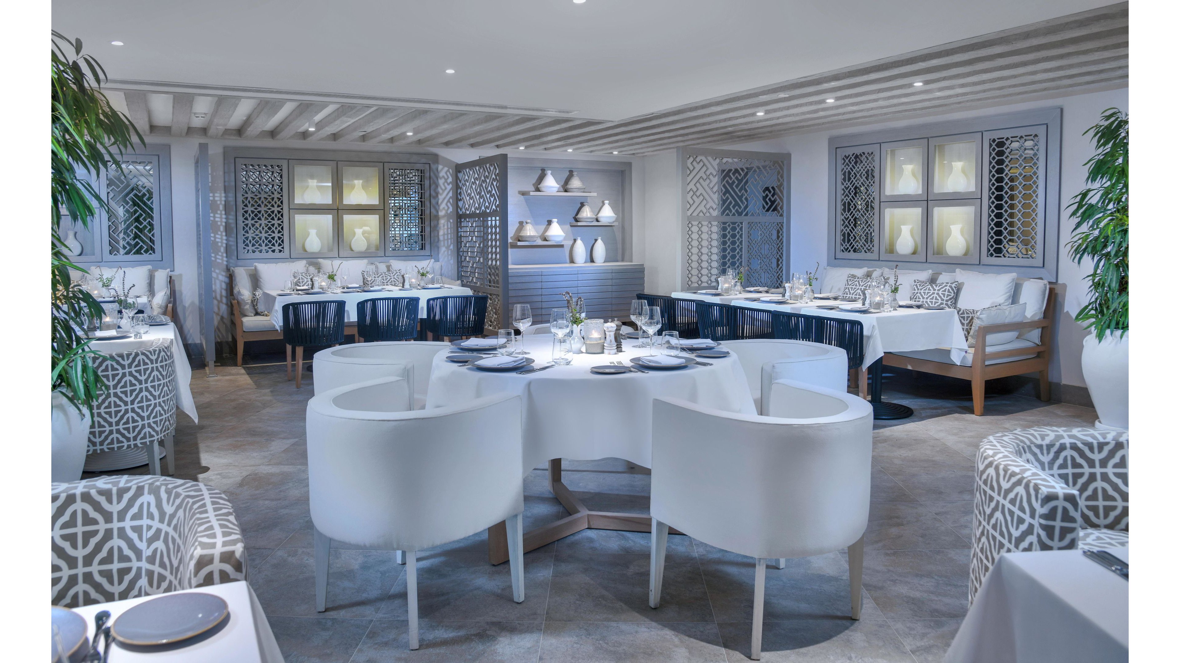 A contemporary restaurant dining area features round tables set with plates and glassware, surrounded by plush white chairs. The setting is bright and airy, with patterned accents and decorative vases displayed on shelves. Soft lighting and neutral tones create a calm, sophisticated atmosphere. No visible people or text are present in the image.