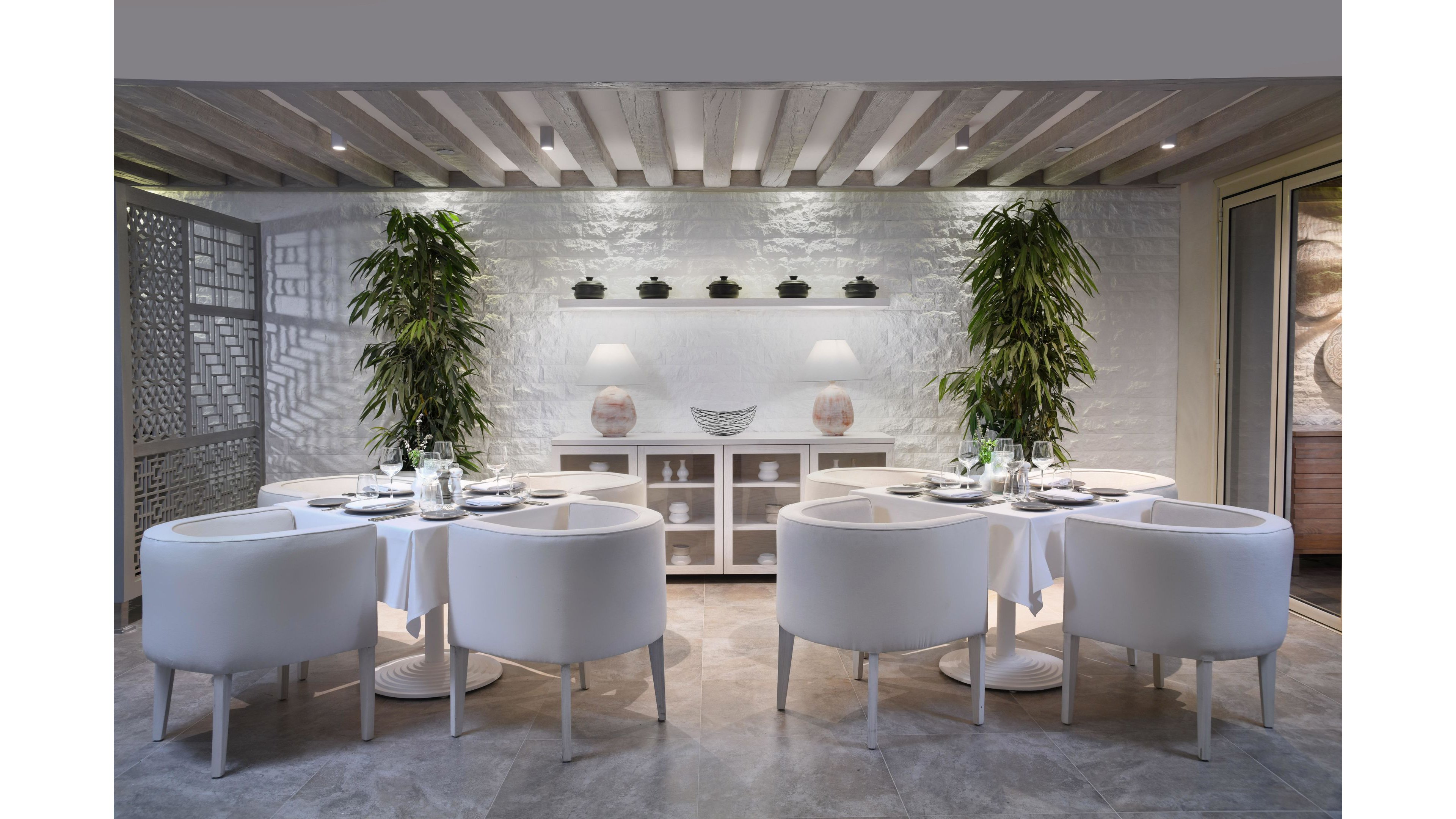 A contemporary restaurant interior features two round tables set for dining, each surrounded by sleek white chairs. Tall green plants frame the tables, adding a touch of nature to the minimalist white and gray palette. The background includes a white brick wall, decorative shelving, and soft lighting from table lamps. The overall mood is calm, inviting, and sophisticated, with a focus on clean lines and modern style.
