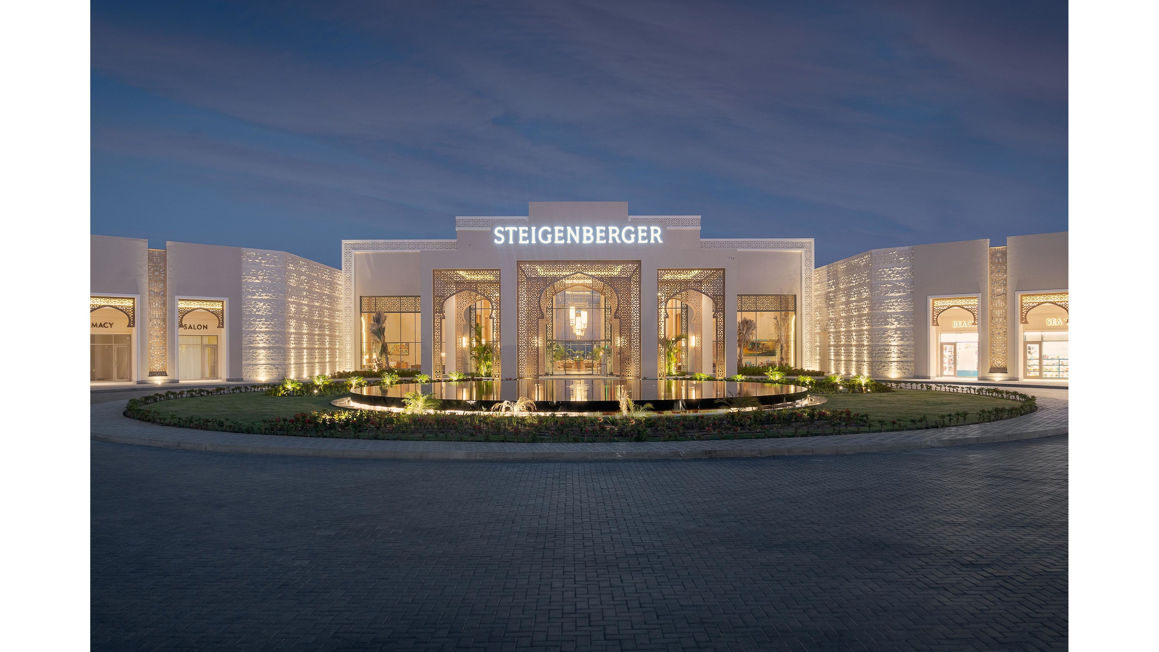 The image showcases the illuminated exterior of a Steigenberger hotel at dusk. The building features elegant architectural details, a grand entrance, and a circular reflecting pool in the foreground. Warm lighting highlights the facade, while the sky transitions to evening blue. The word 'STEIGENBERGER' is clearly visible above the entrance.