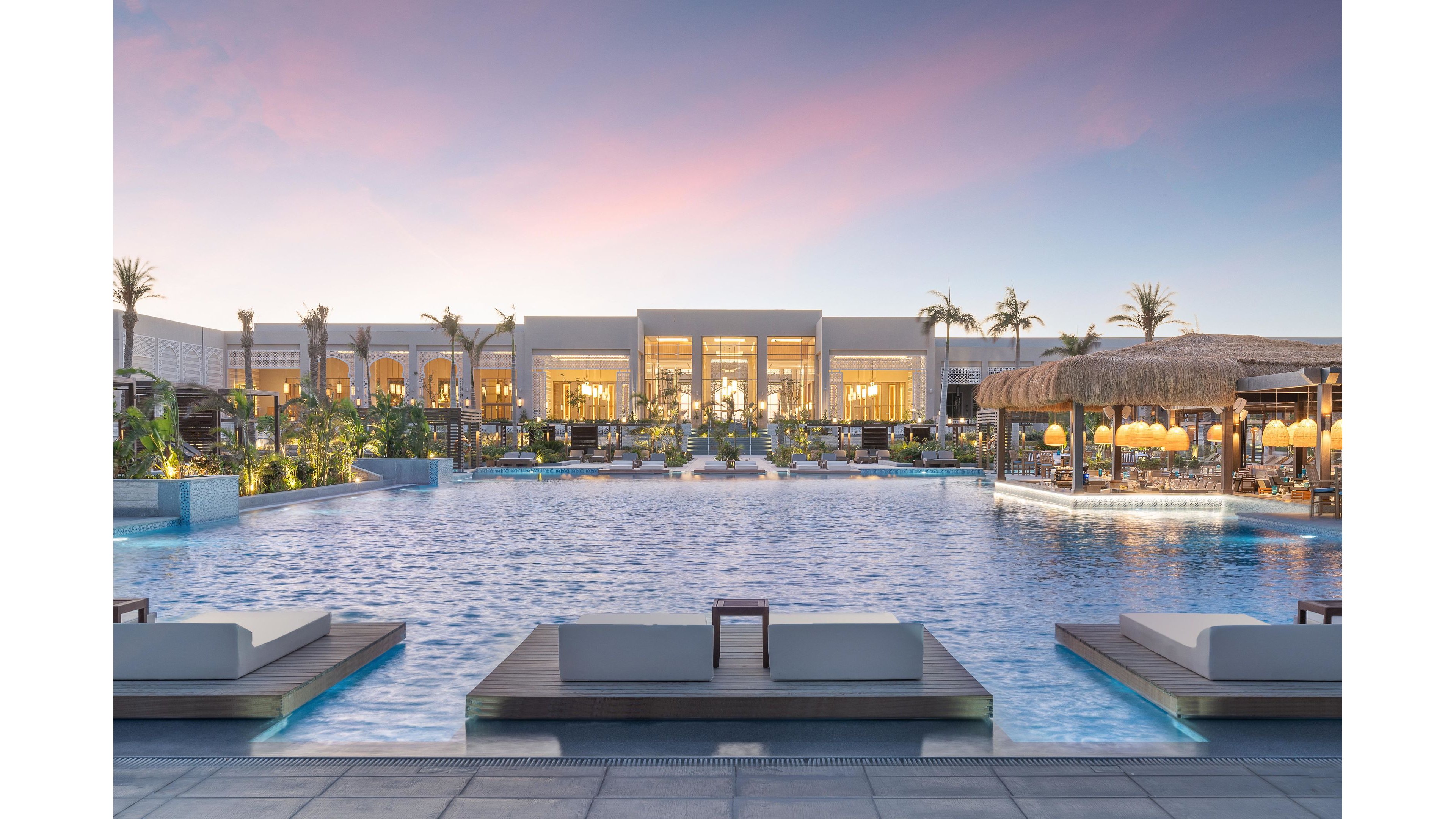 A large, tranquil swimming pool is the centerpiece of a luxury resort, surrounded by modern lounge beds and lush tropical landscaping. The setting is serene, with soft evening light and a pastel sky creating a relaxing atmosphere. Elegant architecture and illuminated outdoor dining areas enhance the upscale ambiance. No visible text or numbers are present in the image.