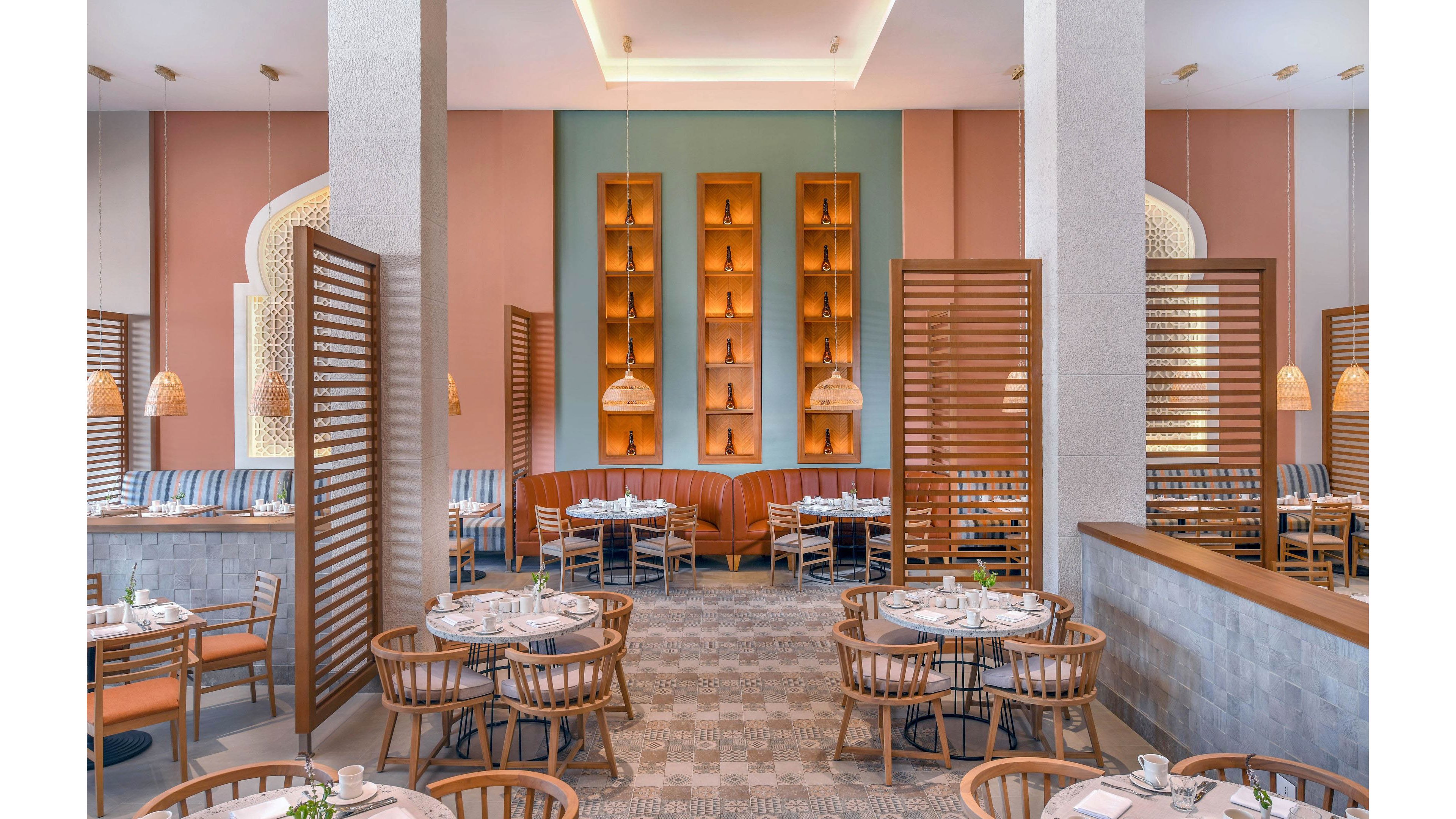 A spacious restaurant dining area features round wooden tables and chairs arranged neatly. The setting includes pastel walls, geometric wooden partitions, and warm pendant lighting, creating a welcoming and contemporary atmosphere. Upholstered seating lines the walls, and decorative alcoves add visual interest. The overall palette is soft with natural wood tones and muted colors.