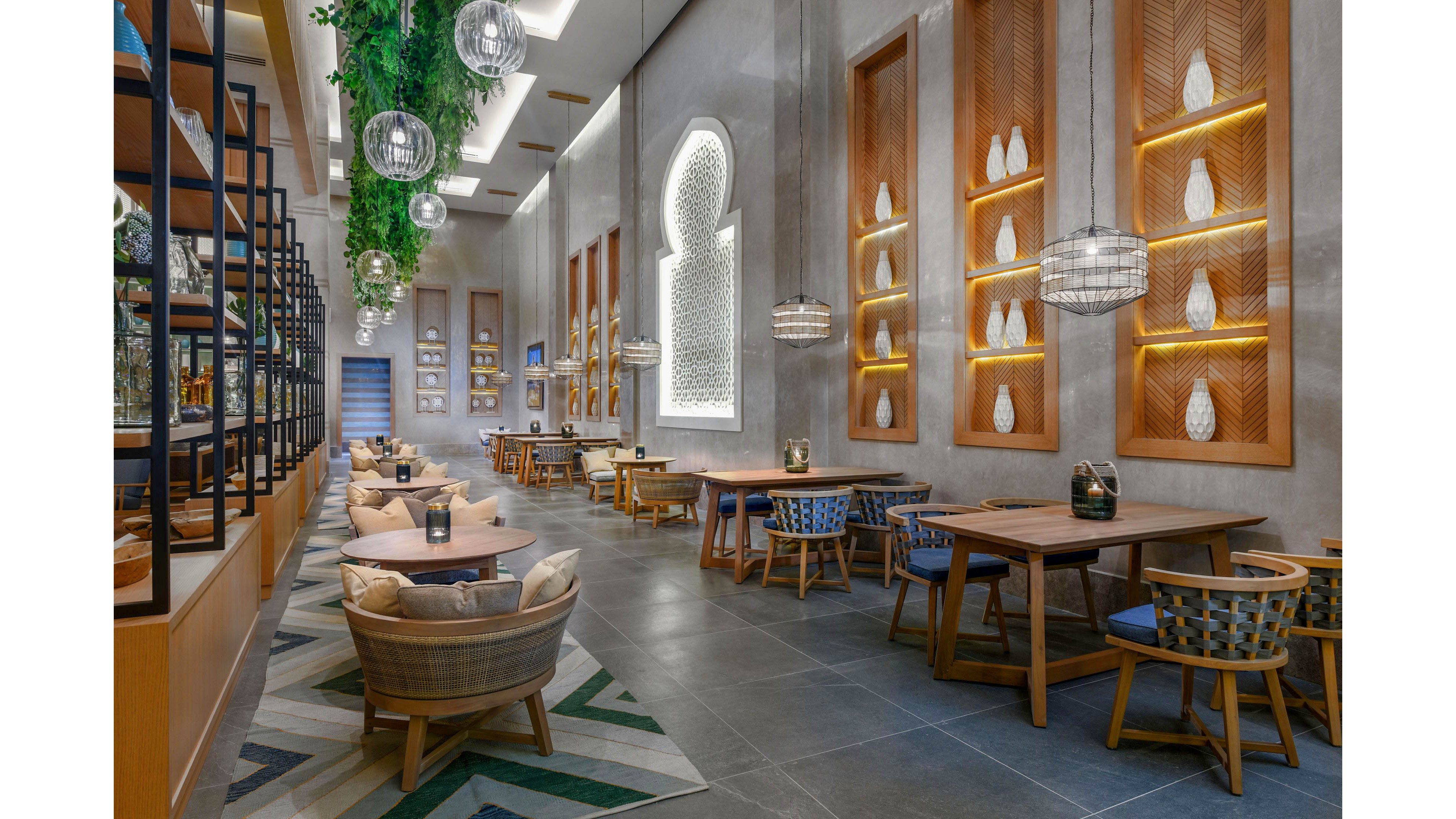 A contemporary restaurant dining area features wooden tables and cushioned chairs arranged along a spacious corridor. The setting includes geometric wall panels with illuminated vases, hanging globe lights, and lush greenery suspended from the ceiling. The color palette is neutral with accents of blue and green, creating a relaxed and inviting atmosphere. No visible people or text are present in the image.