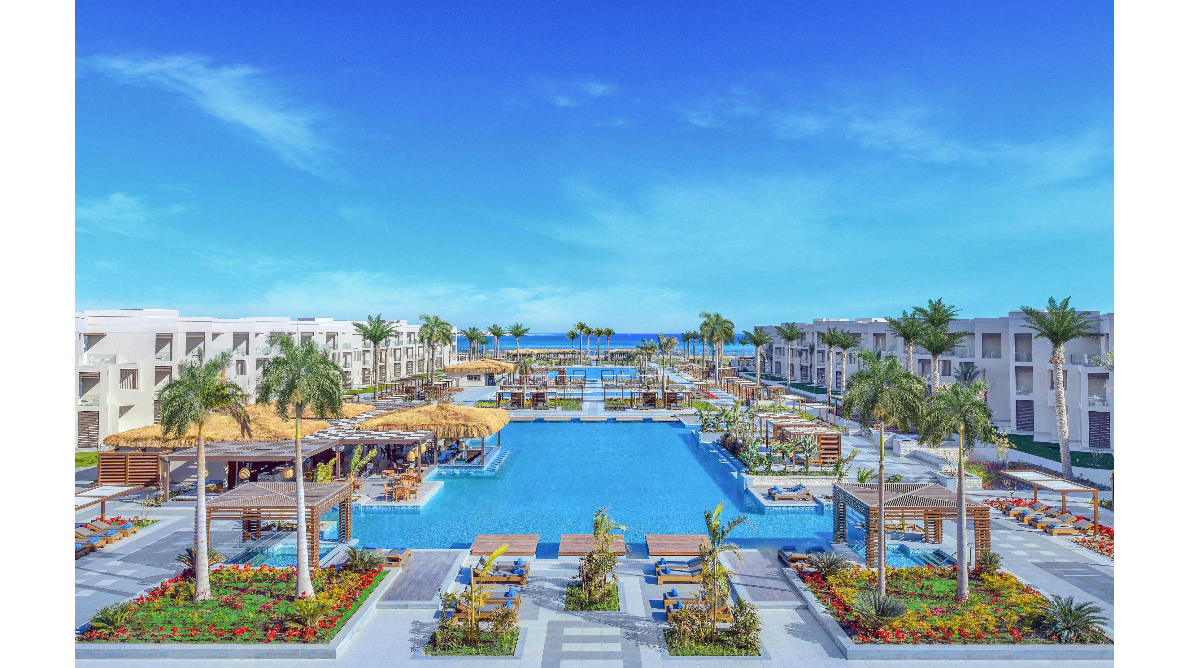 A large, modern resort features a central swimming pool surrounded by palm trees and sun loungers. The setting is outdoors with clear blue skies and the ocean visible in the background. The architecture is contemporary with white buildings and lush landscaping, creating a relaxing and inviting atmosphere. No visible text or numbers are present in the image.