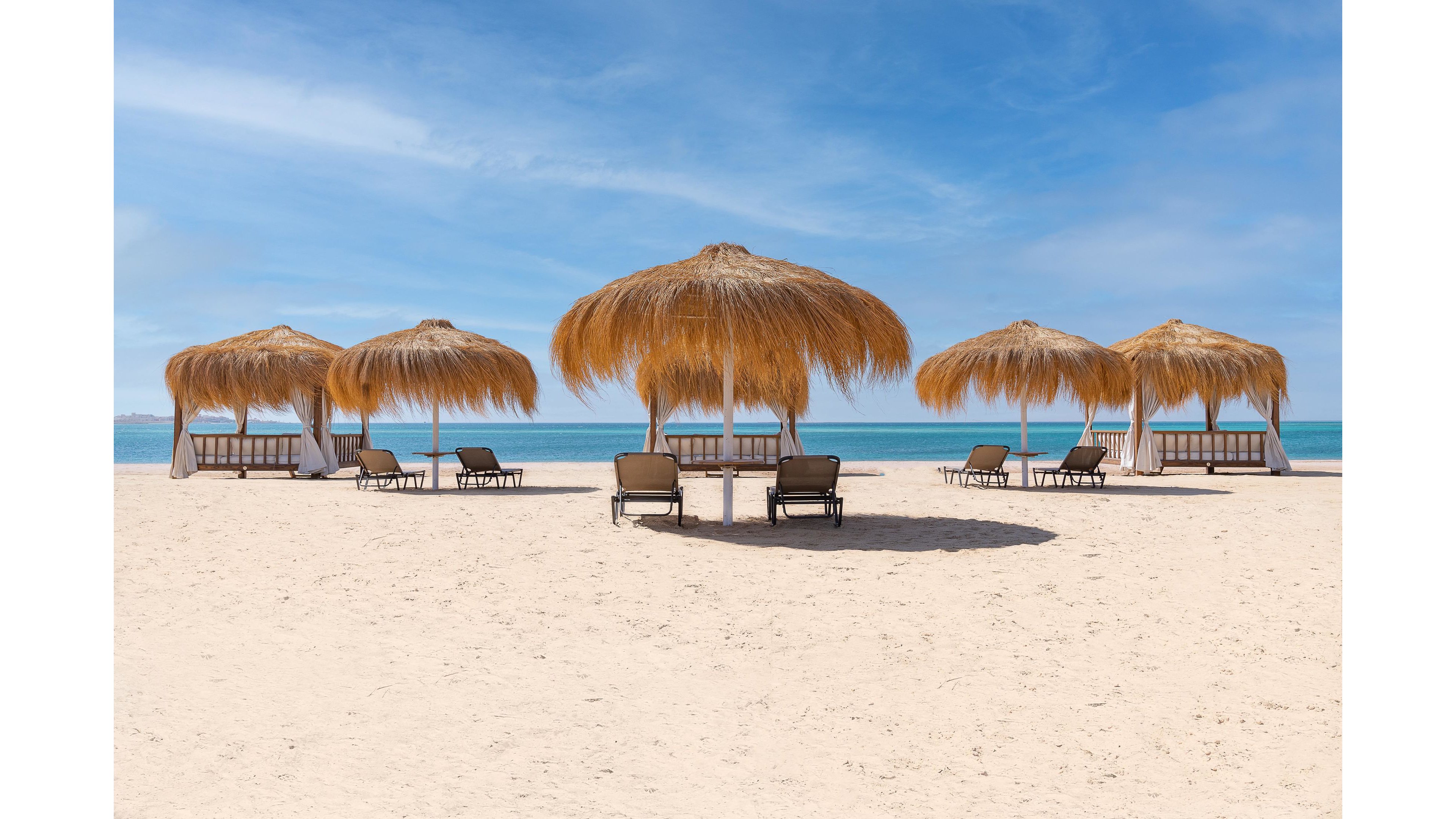 A serene sandy beach features several straw-thatched umbrellas and empty lounge chairs facing the calm blue sea. The setting is tranquil and inviting, with clear skies and turquoise water in the background. The arrangement of loungers under the shade creates a relaxing vacation atmosphere. No people are present, emphasizing peaceful solitude. No visible text or numbers appear in the image.