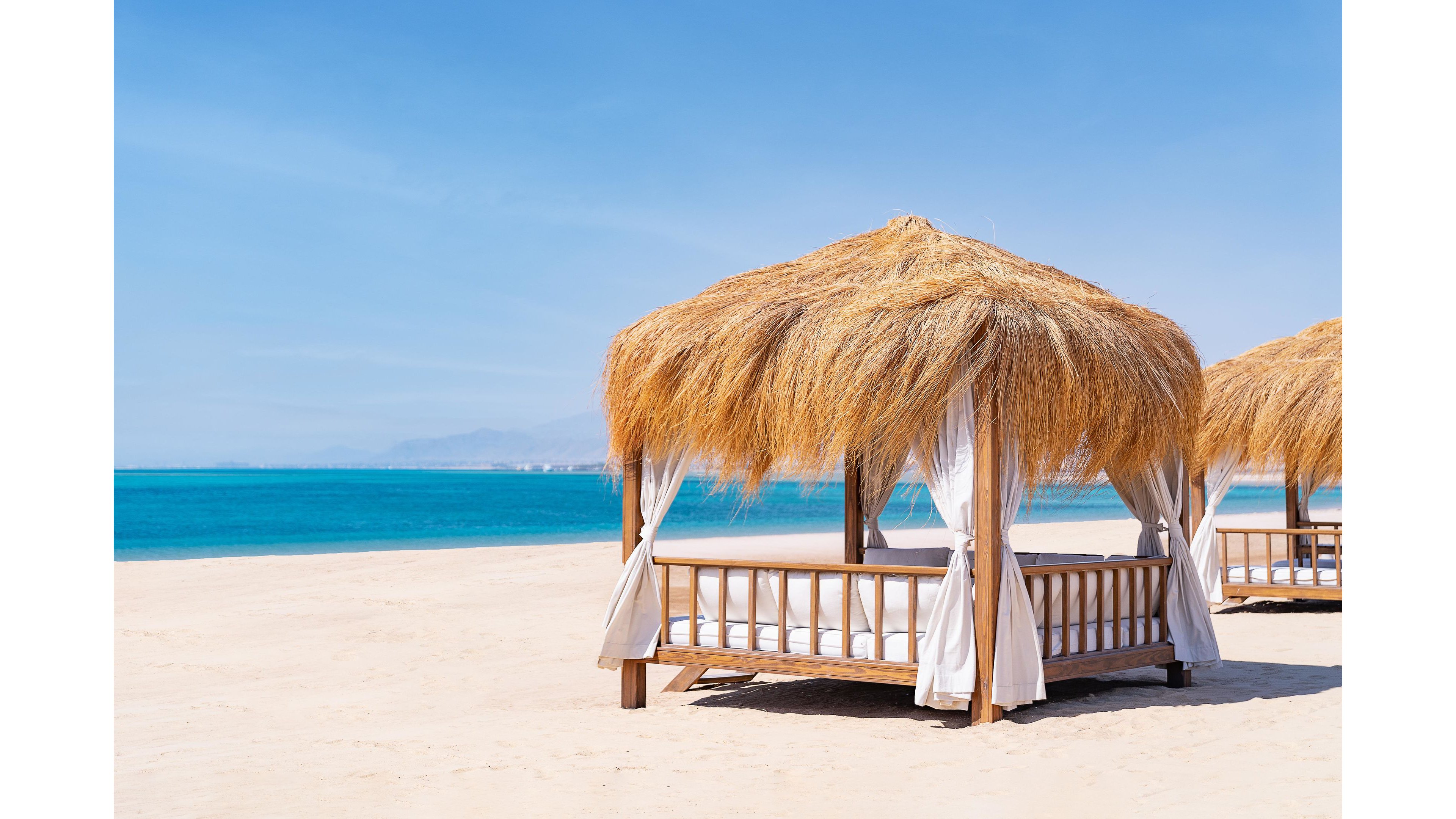 A wooden cabana with a thatched roof stands on a pristine sandy beach, facing a calm turquoise ocean. The cabana is draped with white curtains, creating a relaxing and inviting atmosphere under a clear blue sky. The setting is tranquil and sunlit, evoking a sense of leisure and tropical escape. No people or visible text appear in the image.