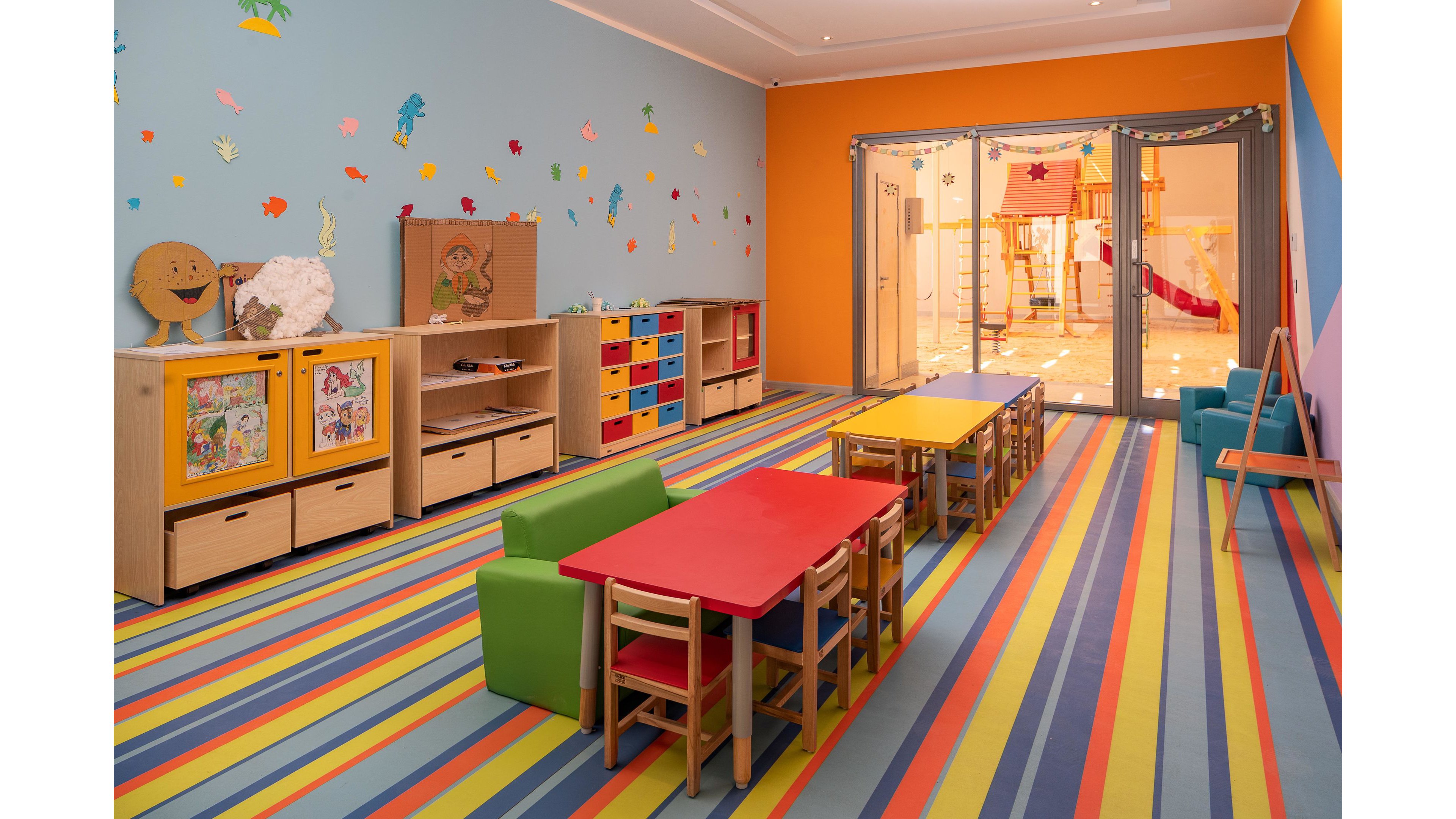 A vibrant preschool classroom features small tables and chairs arranged on a striped, multicolored floor. The walls are decorated with playful cutouts and art, while shelves hold toys and educational materials. Large glass doors reveal an adjacent indoor play area with slides. The setting is bright and inviting, designed for young children. No visible text or numbers are present in the image.