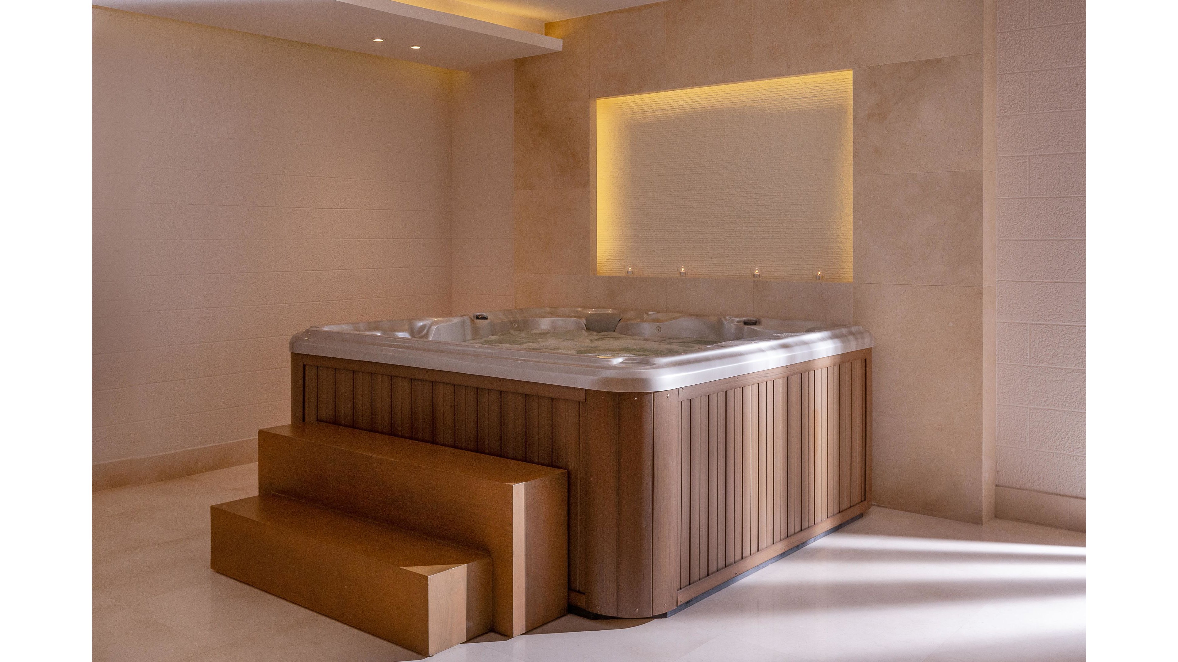 A contemporary hot tub with sleek wooden paneling is featured in a minimalist indoor spa setting. The tub is accompanied by matching wooden steps for easy access and is illuminated by soft, recessed lighting and a glowing wall accent. The neutral color palette and clean lines create a relaxing and luxurious atmosphere. No visible text, logos, or numeric values are present in the image.