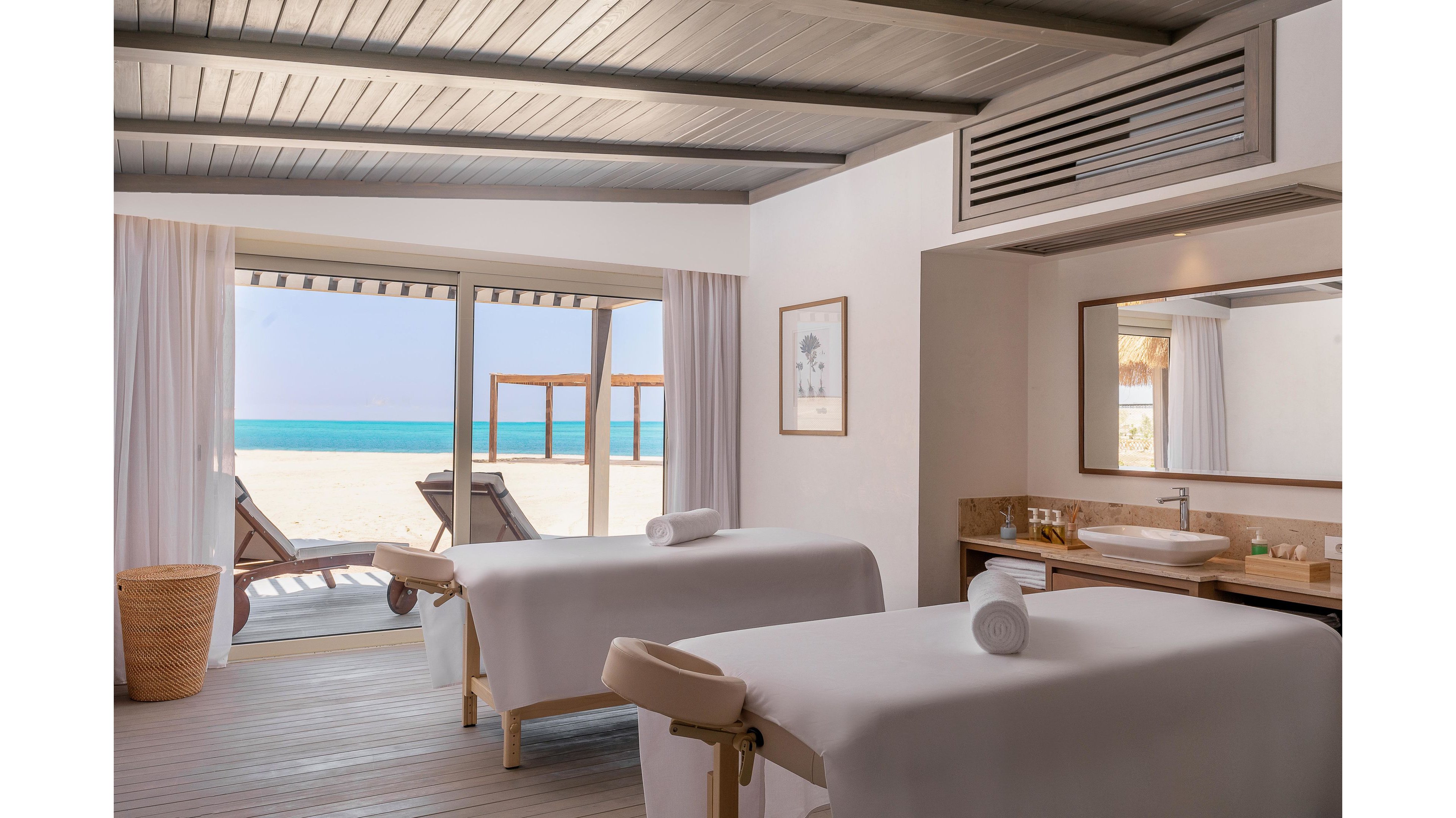 A serene spa treatment room features two massage tables facing large windows that open to a sandy beach and turquoise ocean. The interior is decorated in neutral tones with natural light streaming in, creating a calm and relaxing atmosphere. Minimalist decor includes a framed artwork, a large mirror, and a modern sink area. Outside, wooden lounge chairs and cabanas are visible on the beach.