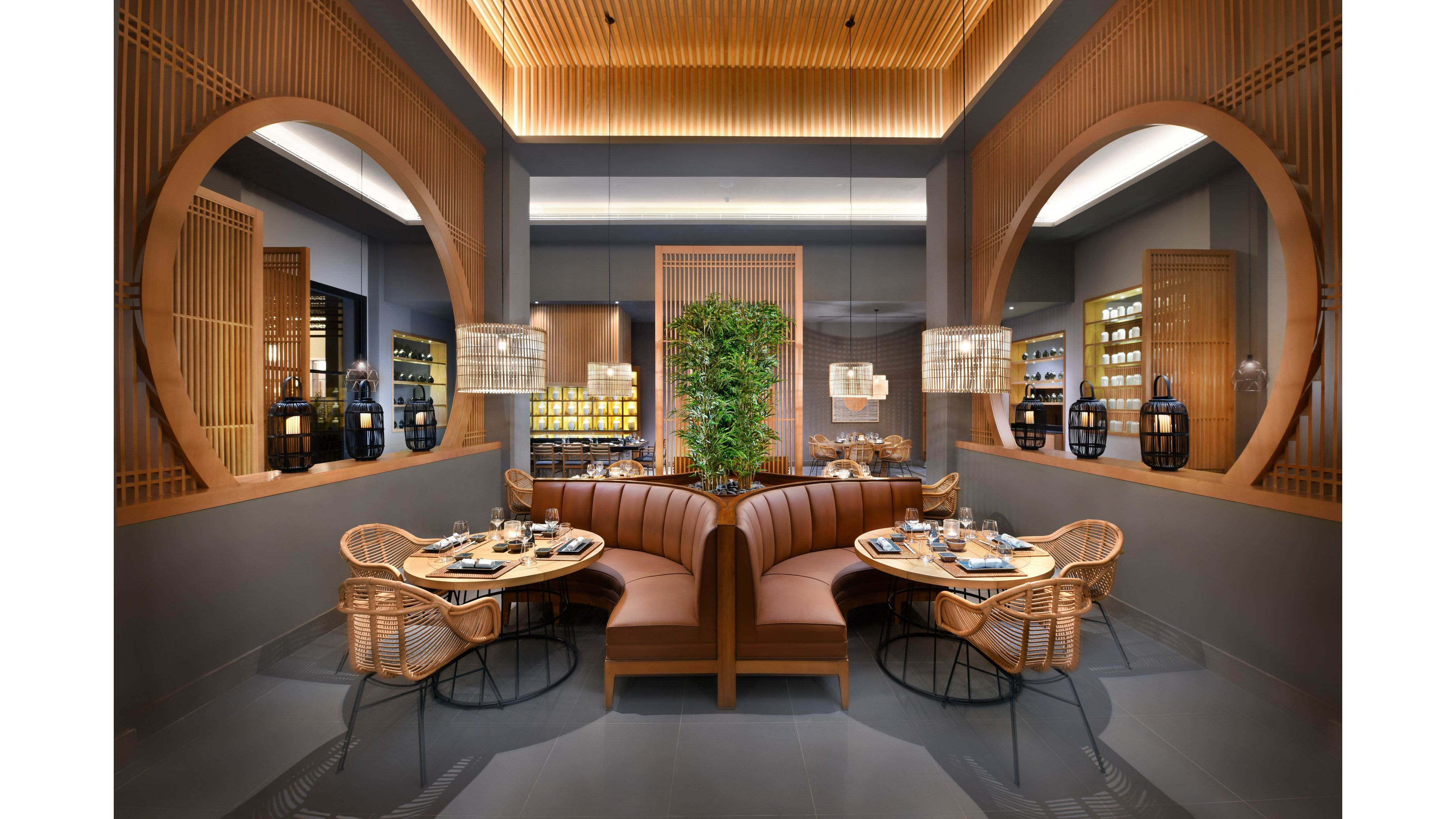 A contemporary restaurant dining area features elegant wooden paneling and circular architectural details. The setting includes cozy brown leather booths, rattan chairs, and neatly set tables, creating a welcoming atmosphere. Warm lighting and a central green plant add to the tranquil, sophisticated mood. Decorative shelving and glassware are visible in the background, enhancing the upscale ambiance.