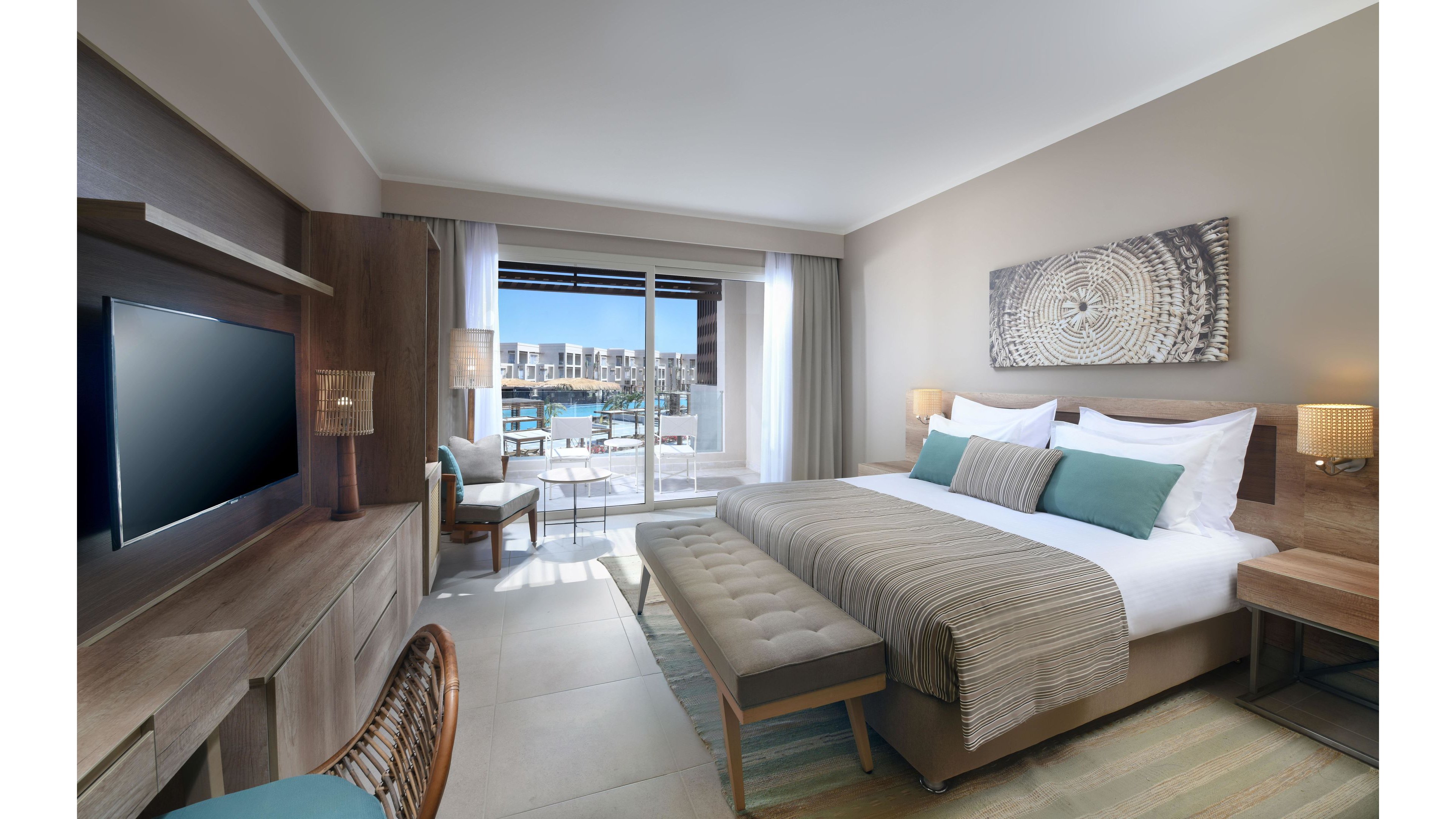 A spacious, contemporary hotel room features a large bed with striped bedding and teal accent pillows. The room includes a flat-screen TV, wooden furniture, and a decorative wall art piece above the bed. Natural light fills the space through sliding glass doors leading to a balcony overlooking a waterfront or pool area. The neutral color palette and soft lighting create a calm, inviting atmosphere. No visible text or numbers are present in the image.