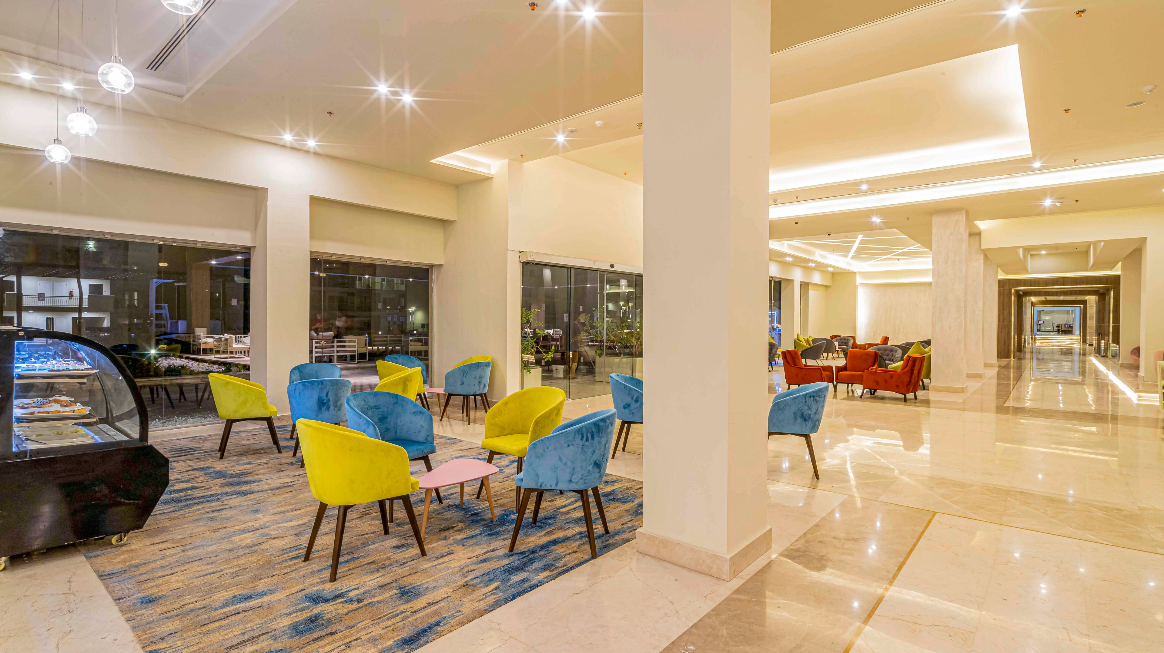 Spacious modern hotel lobby lounge featuring bright blue and yellow chairs around small tables. Large windows line the wall, reflecting the interior lighting and hinting at an outdoor view. A display case stands to the side, suggesting a café or dessert area within the lobby. Warm recessed lighting and polished floors create an inviting, contemporary atmosphere extending down a long corridor.