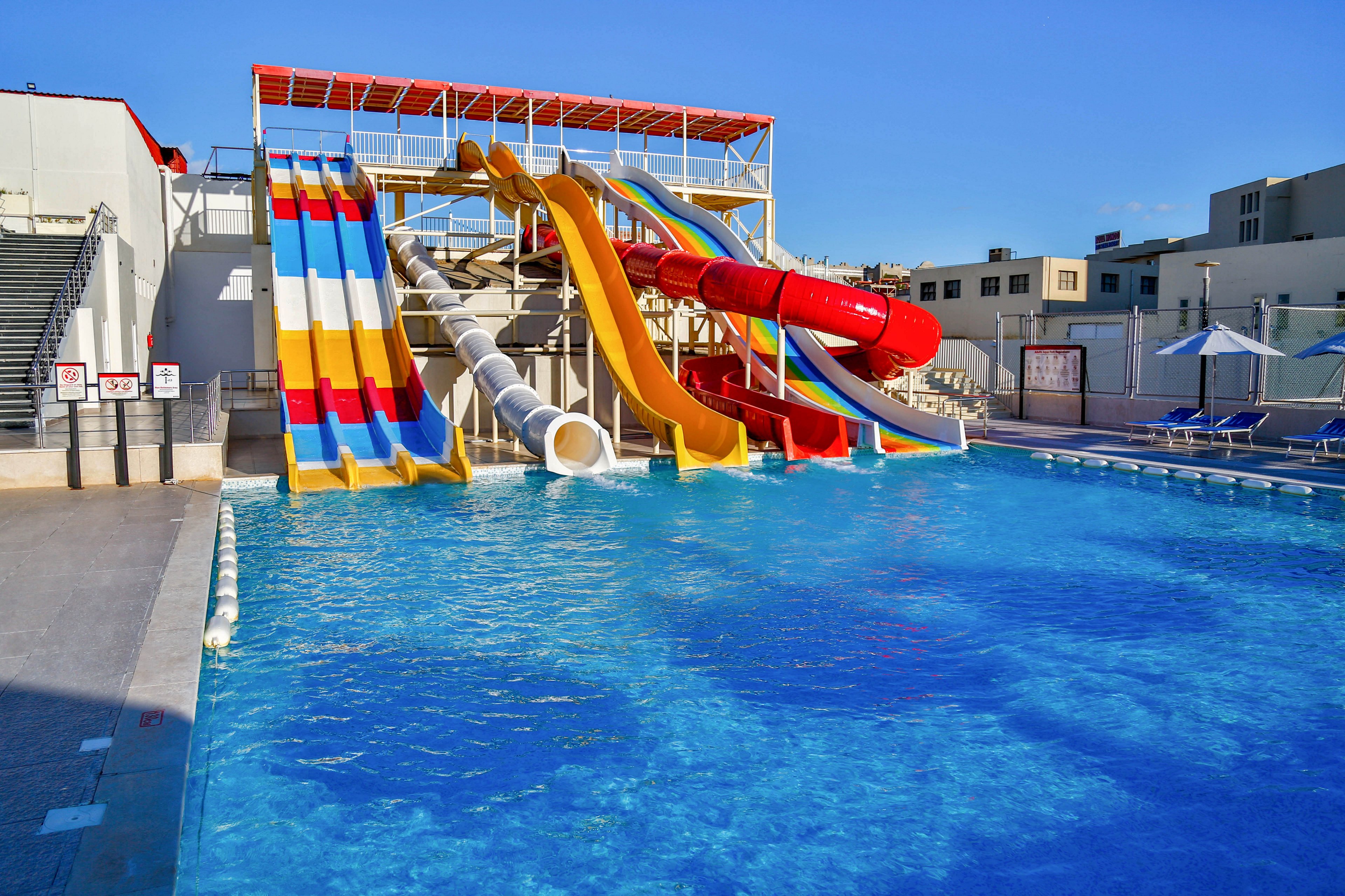 A group of large, colorful water slides empties into a bright blue outdoor swimming pool. The slides include open and enclosed tubes in red, yellow, white, and striped designs. The surrounding area features poolside seating, umbrellas, and nearby buildings under a clear sky. The scene suggests a modern water park or recreational facility ready for visitors.