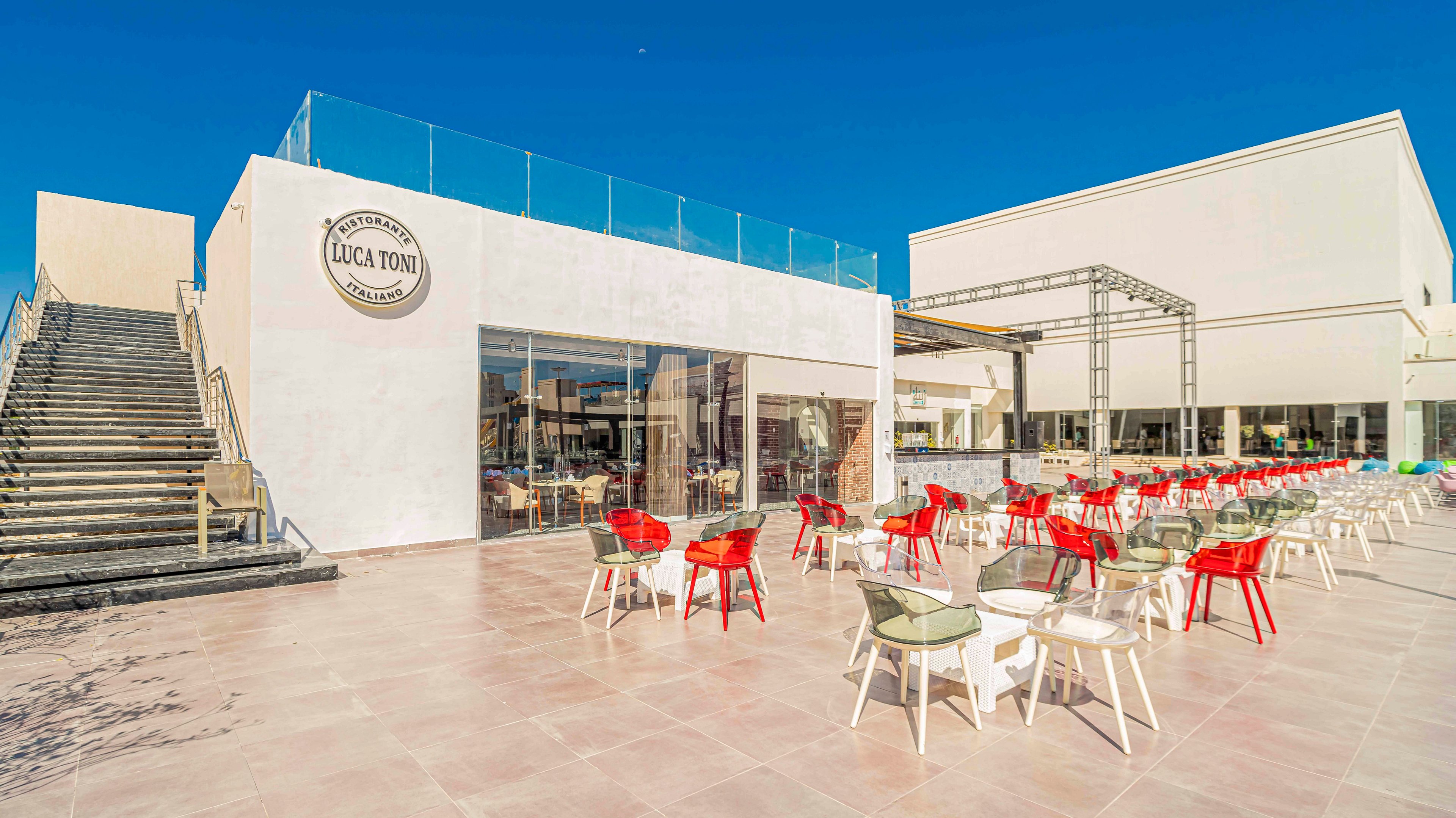 A contemporary white building houses a café with large glass windows and an expansive outdoor terrace. The name "LUCA TONI" appears on the façade in a circular sign. Numerous red and white chairs surround glass-topped tables arranged across the open patio. A wide staircase leads to an upper level, and the scene is set under a clear blue sky, suggesting a bright, sunny day.