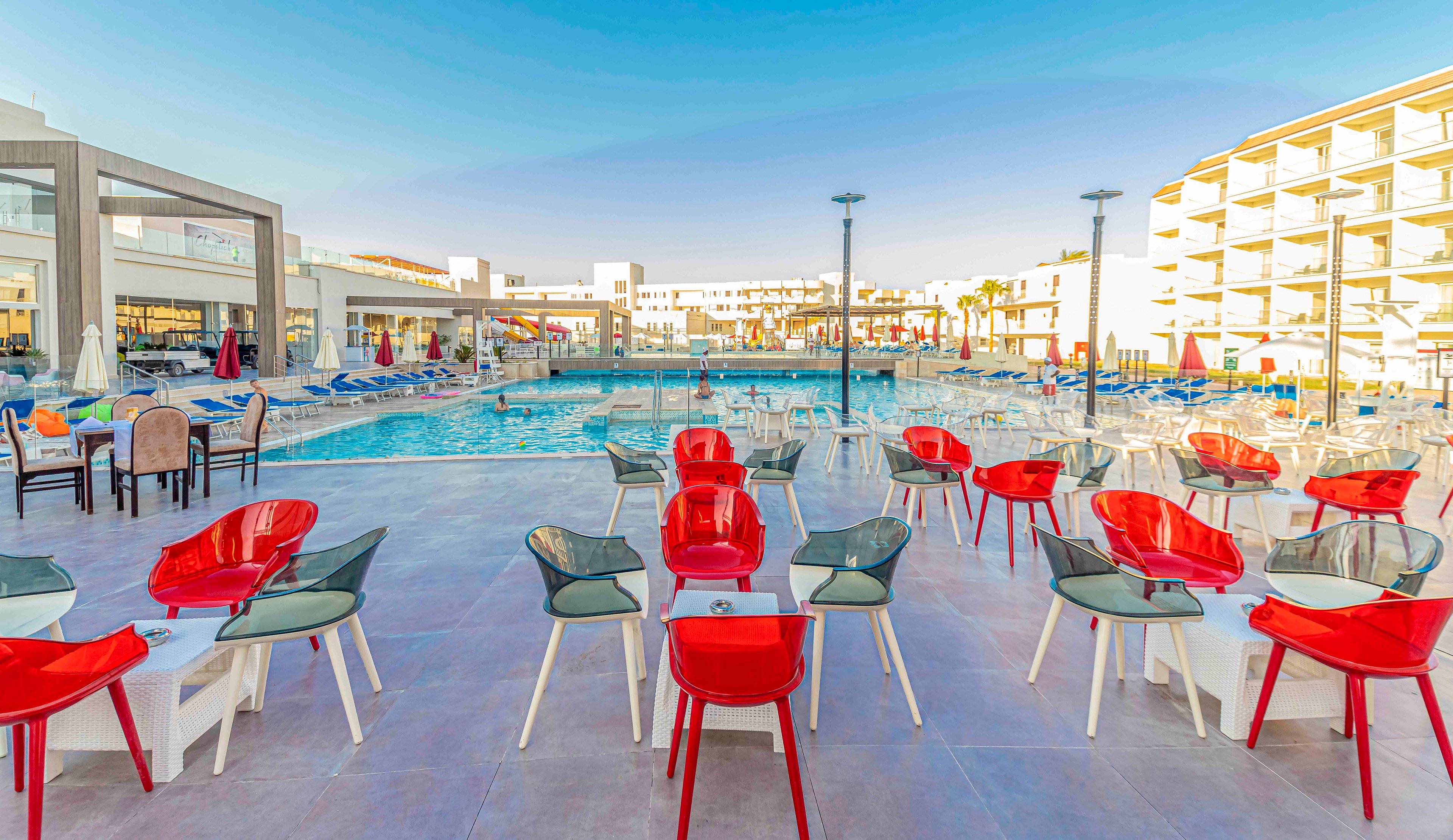 An outdoor resort pool area features rows of modern red and transparent chairs arranged on a spacious terrace. In the background, a large swimming pool is surrounded by blue sun loungers and umbrellas. Multi-story hotel buildings frame the scene under a clear blue sky. The bright colors and clean architecture create a relaxed vacation atmosphere.