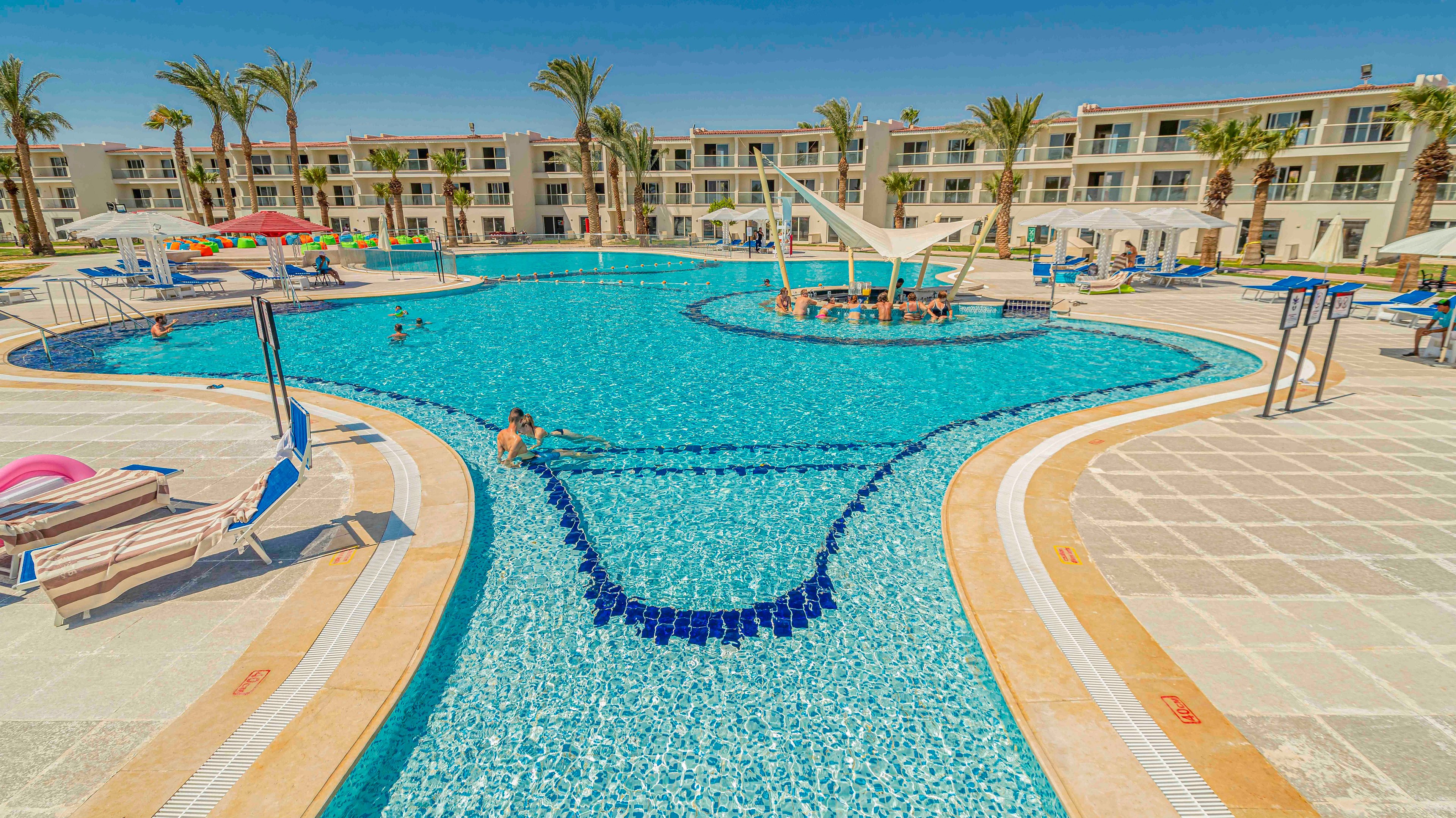 A large outdoor swimming pool is surrounded by sun loungers and umbrellas at a modern resort. Low-rise hotel buildings with balconies and palm trees frame the bright blue pool. A few people relax in the water and on the deck under clear, sunny skies. The scene conveys a warm vacation atmosphere with clean lines and vibrant colors.