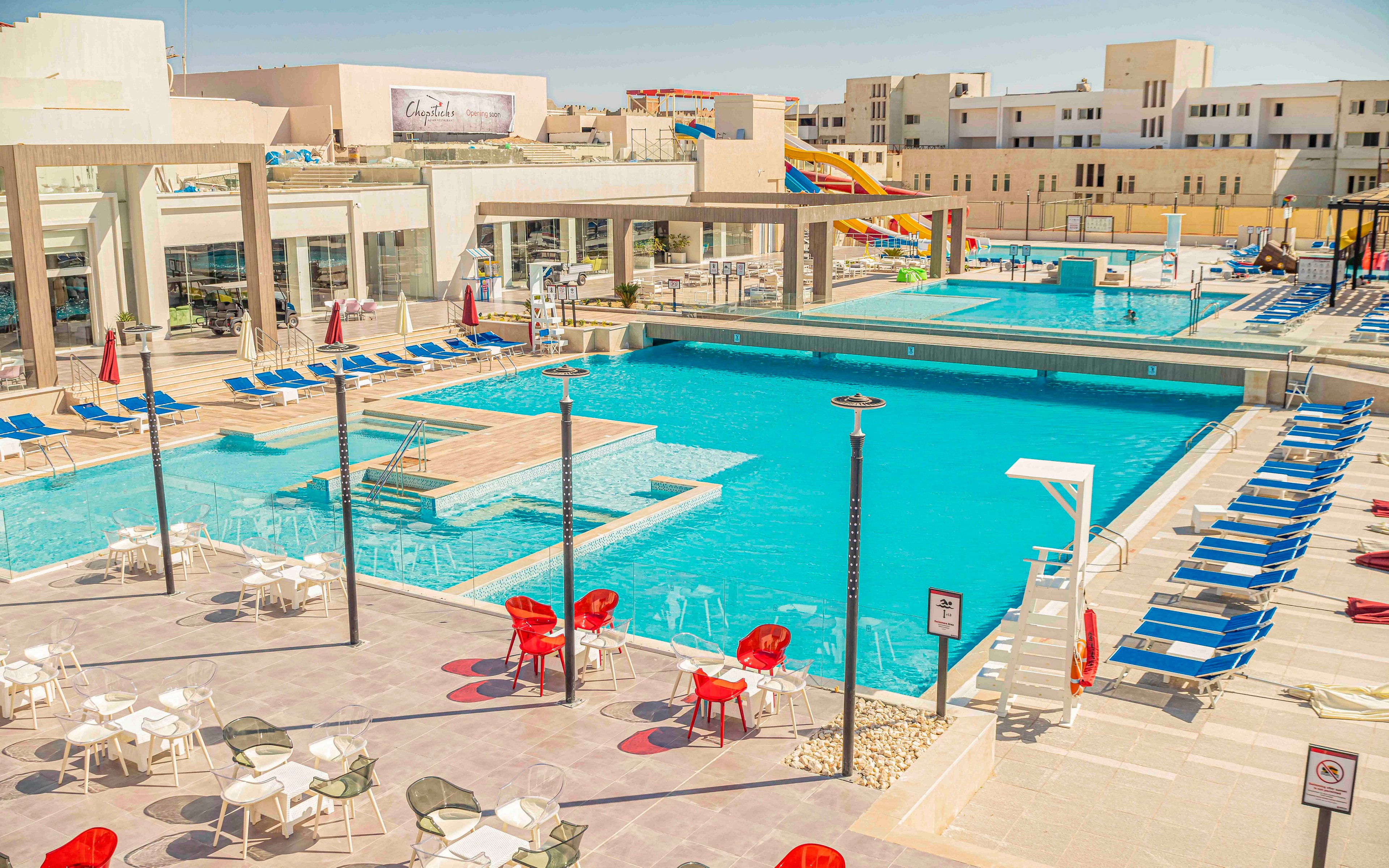 A large outdoor swimming pool complex is shown under bright sunlight at a modern resort. Multiple pools with clear blue water are surrounded by rows of blue lounge chairs and umbrellas. White and red patio chairs and tables occupy the foreground terrace, while water slides and additional pools appear in the background. The scene is empty of people, emphasizing a clean, relaxing vacation atmosphere.
