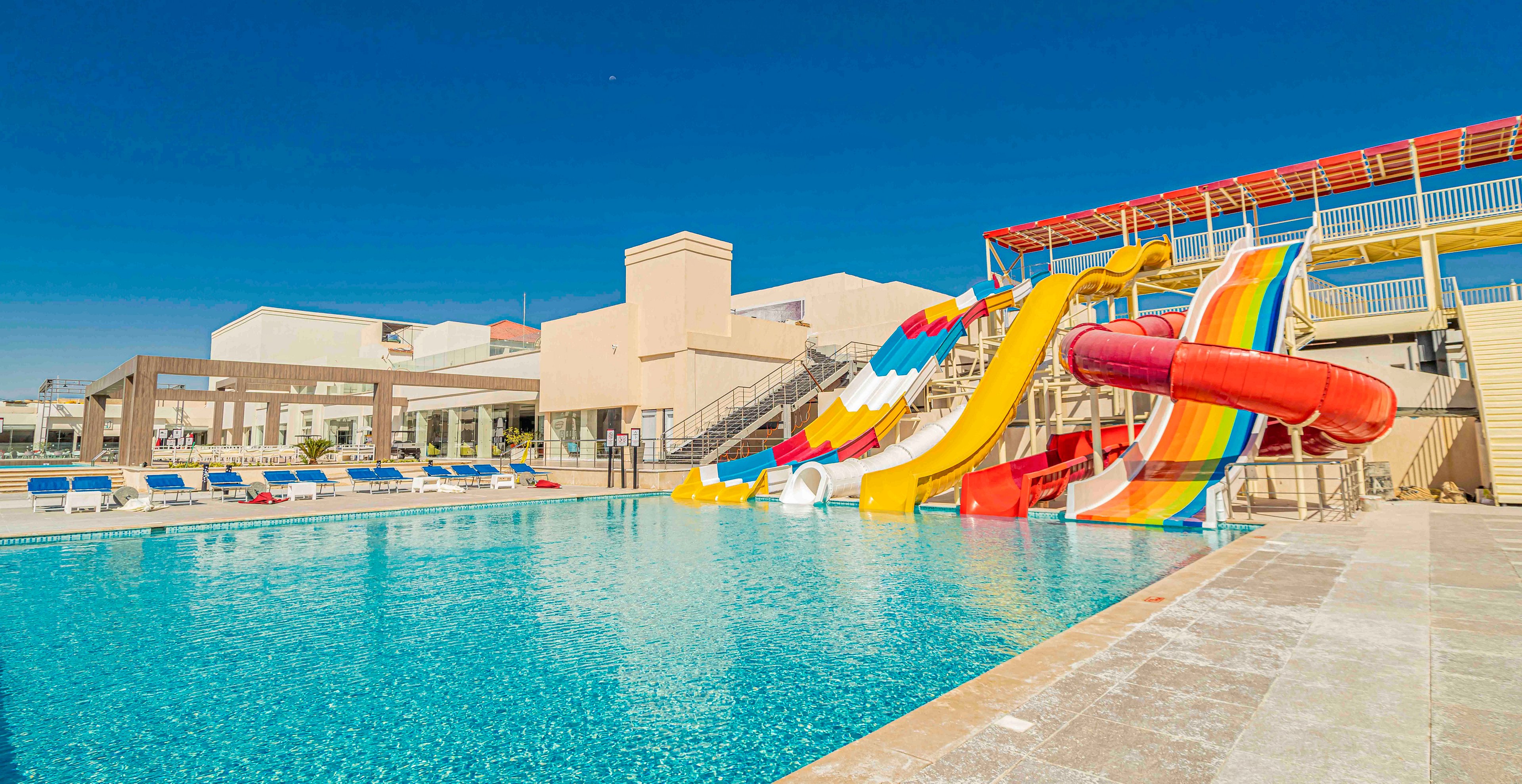 An outdoor resort swimming pool is shown under a clear blue sky, featuring several colorful water slides empty of people. The slides include straight and curved designs in bright red, yellow, orange, and white, leading directly into the pool. Sun loungers with blue cushions line the far side of the pool near modern low-rise buildings. The scene suggests a warm, sunny day ideal for family vacations and leisure activities.