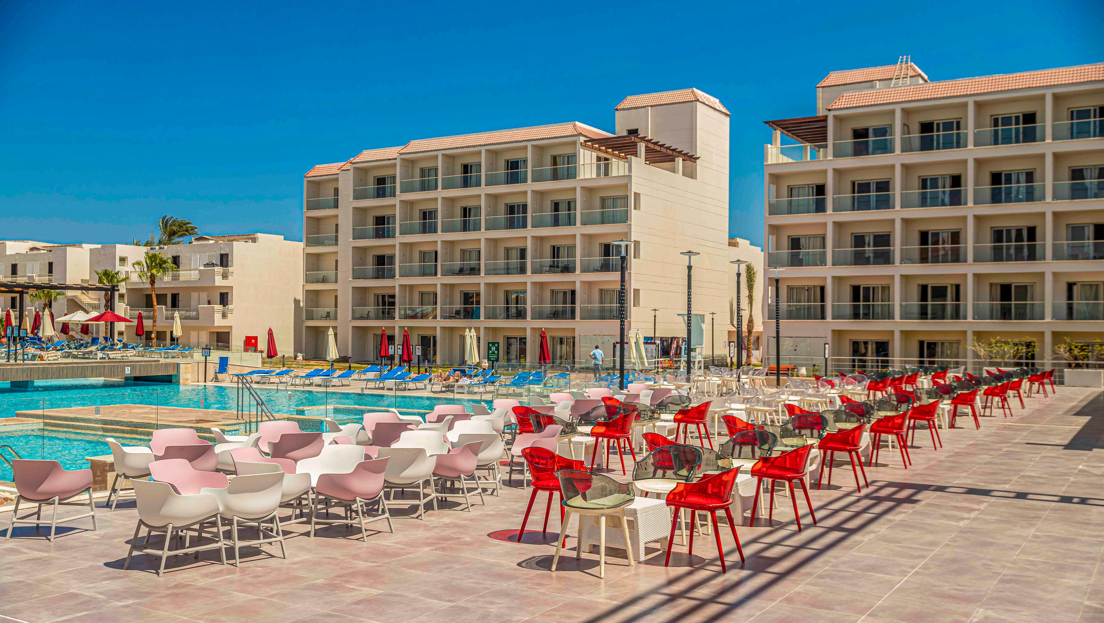 A contemporary resort hotel features multiple mid-rise buildings surrounding a large outdoor swimming pool. Rows of red and white chairs and tables are arranged on a spacious poolside terrace. Sun loungers and umbrellas line the pool, suggesting a relaxing vacation setting. The bright blue sky and strong sunlight create a warm, summery atmosphere.