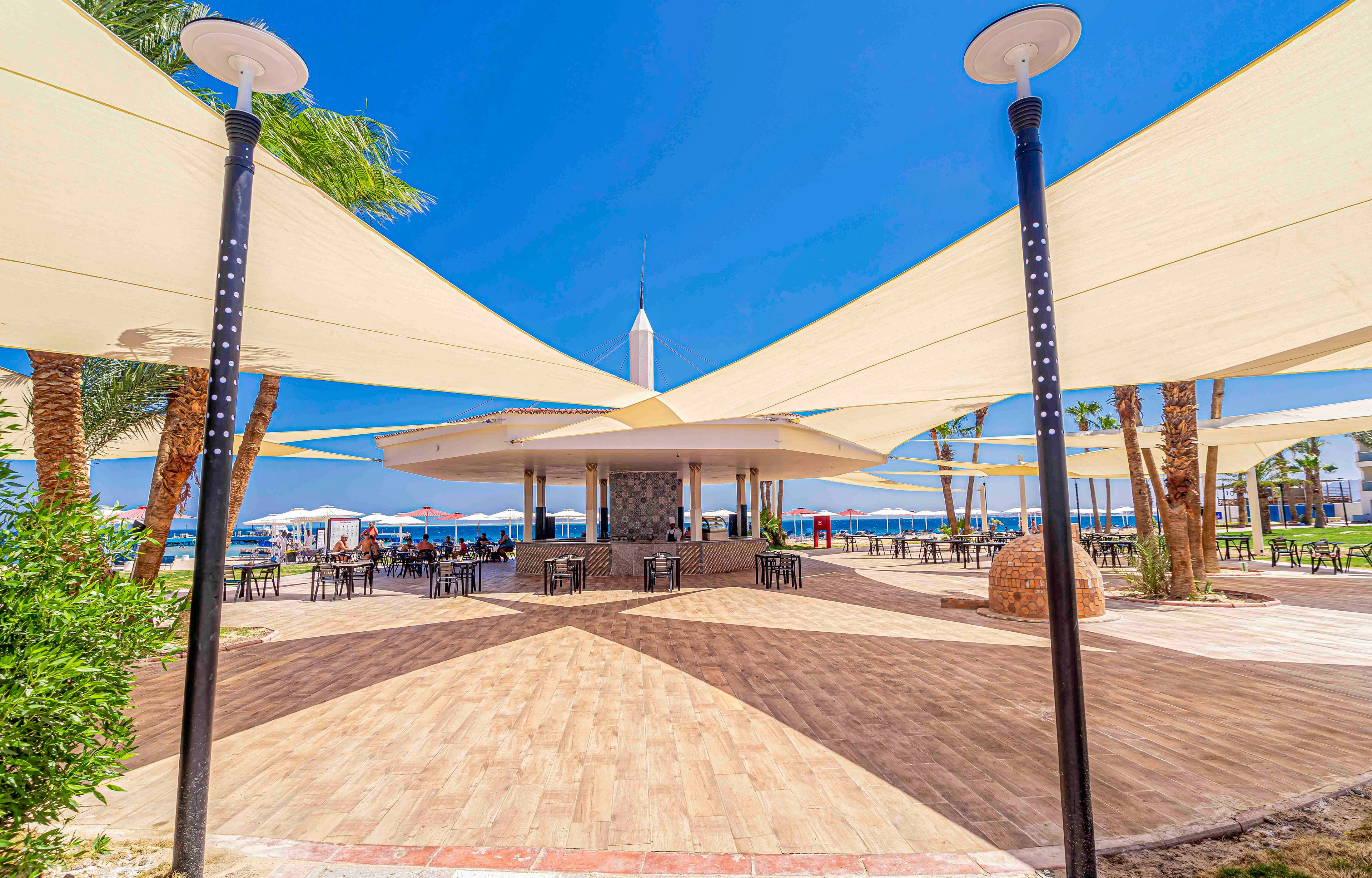 An open-air beachfront cafe is shaded by large triangular fabric sails stretched between tall black poles. The seating area features scattered tables and chairs on a wide tiled terrace. Palm trees surround the space, and the sea is visible in the background under a clear blue sky. The bright sunlight and vivid colors create a relaxed, summery vacation atmosphere.