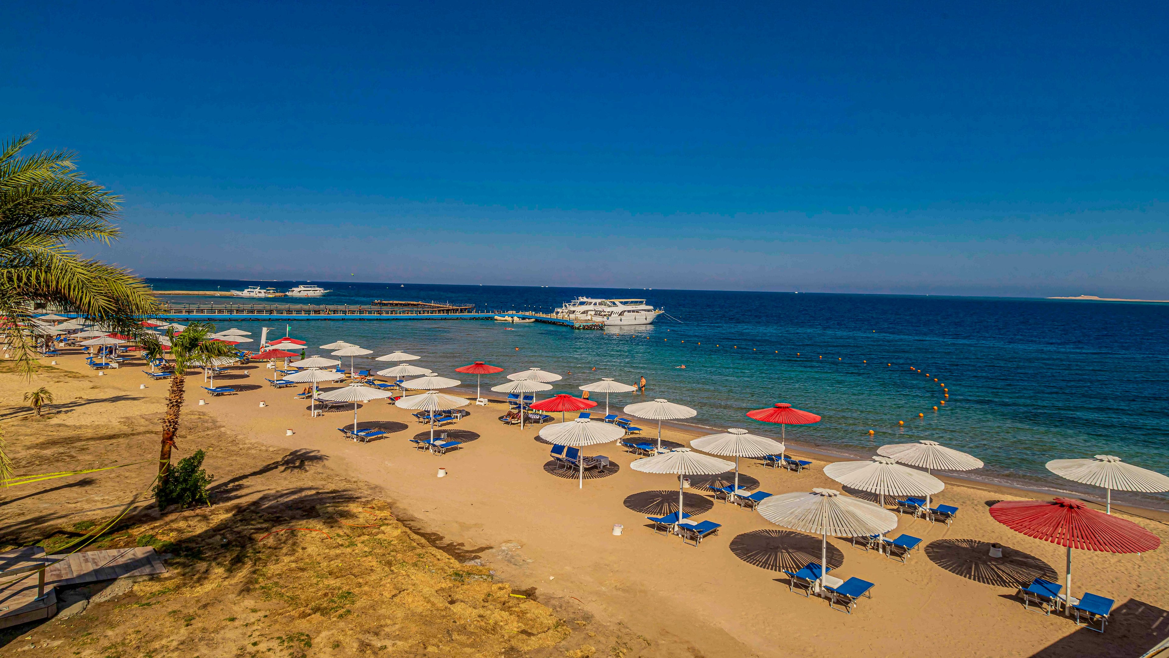 A sandy resort beach is lined with rows of colorful sun umbrellas and lounge chairs facing the sea. Calm blue water stretches to the horizon under a clear sky, with a large white boat anchored offshore. A palm tree and walkway frame the foreground, emphasizing the organized leisure setting. The scene suggests a warm, relaxing vacation destination with ample space for sunbathing and swimming.