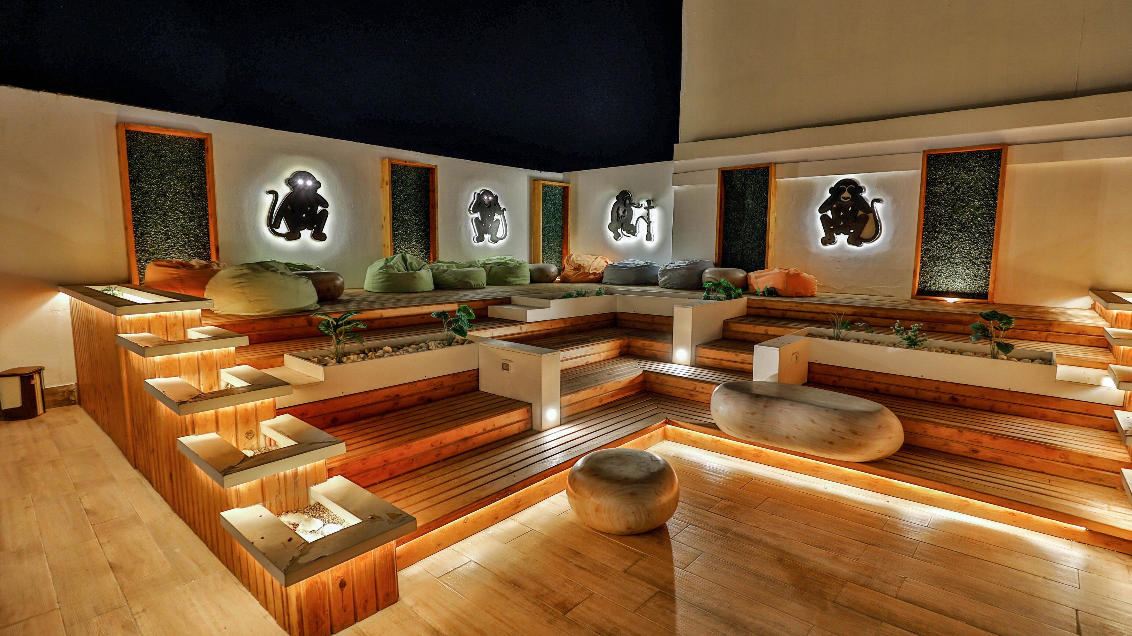 A contemporary rooftop lounge features tiered wooden seating with integrated lighting and smooth stone accents. Soft cushions are arranged along the back wall, creating a relaxed and informal seating area. Illuminated wall art panels with stylized figures add a decorative focal point to the space. Warm ambient lighting highlights the steps, planters, and architectural details against the dark night sky.