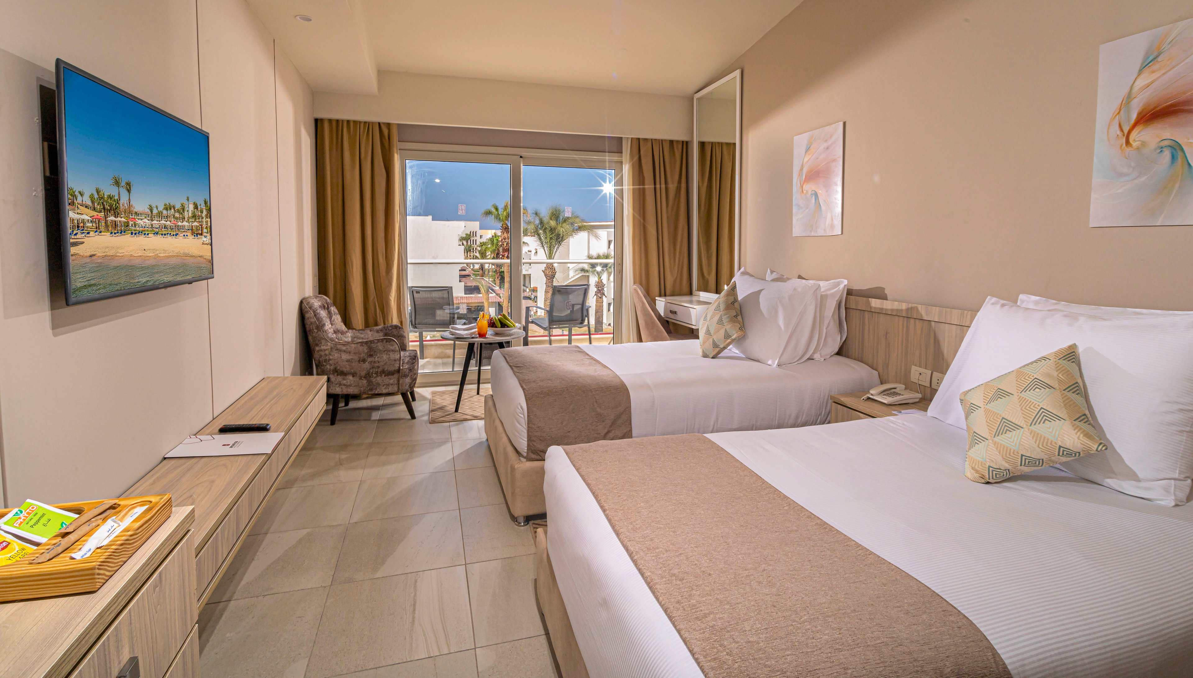 Contemporary hotel room featuring two neatly made twin beds with neutral bedding and accent pillows. A wall-mounted flat-screen television faces the beds, and a desk with a chair sits along the opposite wall. Large sliding glass doors open to a balcony with outdoor seating and a view of palm trees and resort buildings. Warm lighting, beige tones, and minimalist artwork create a calm, inviting atmosphere suitable for vacation or business travel.