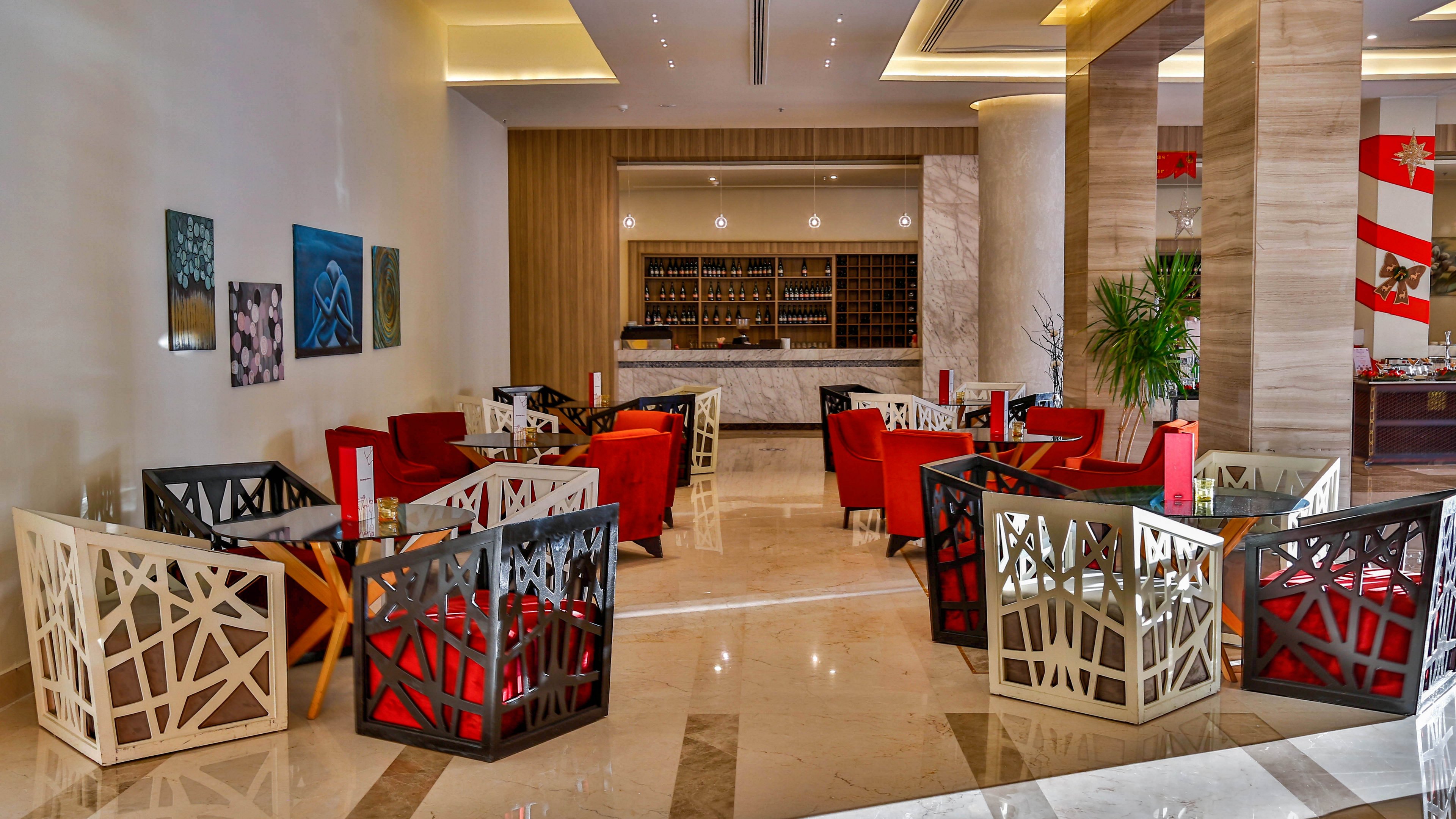 Contemporary lounge area features geometric armchairs arranged around small tables in a spacious hotel lobby. A sleek bar with bottles and glassware is visible in the background, framed by warm wooden paneling. Polished marble floors, tall columns, and recessed ceiling lighting create a bright, upscale atmosphere. Red and neutral tones, abstract wall art, and indoor plants add color and visual interest to the scene.