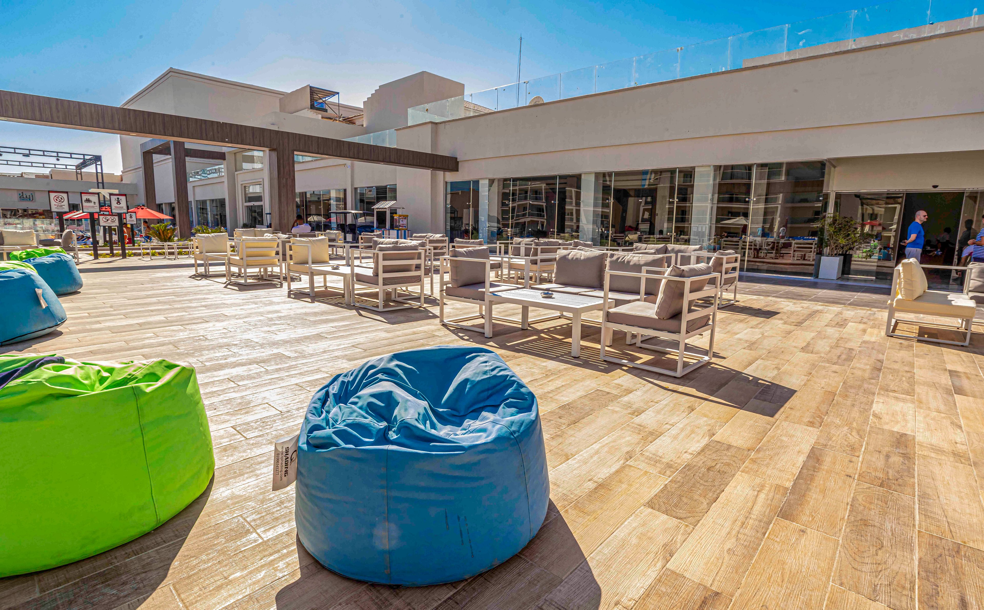 A spacious outdoor terrace features colorful beanbag chairs and modern lounge seating under a bright blue sky. The area is paved with wood-look flooring and bordered by a contemporary building with large glass windows. White sofas and tables are arranged for socializing or relaxing in the sun. The scene suggests a casual, resort-like atmosphere ideal for leisure and gatherings.