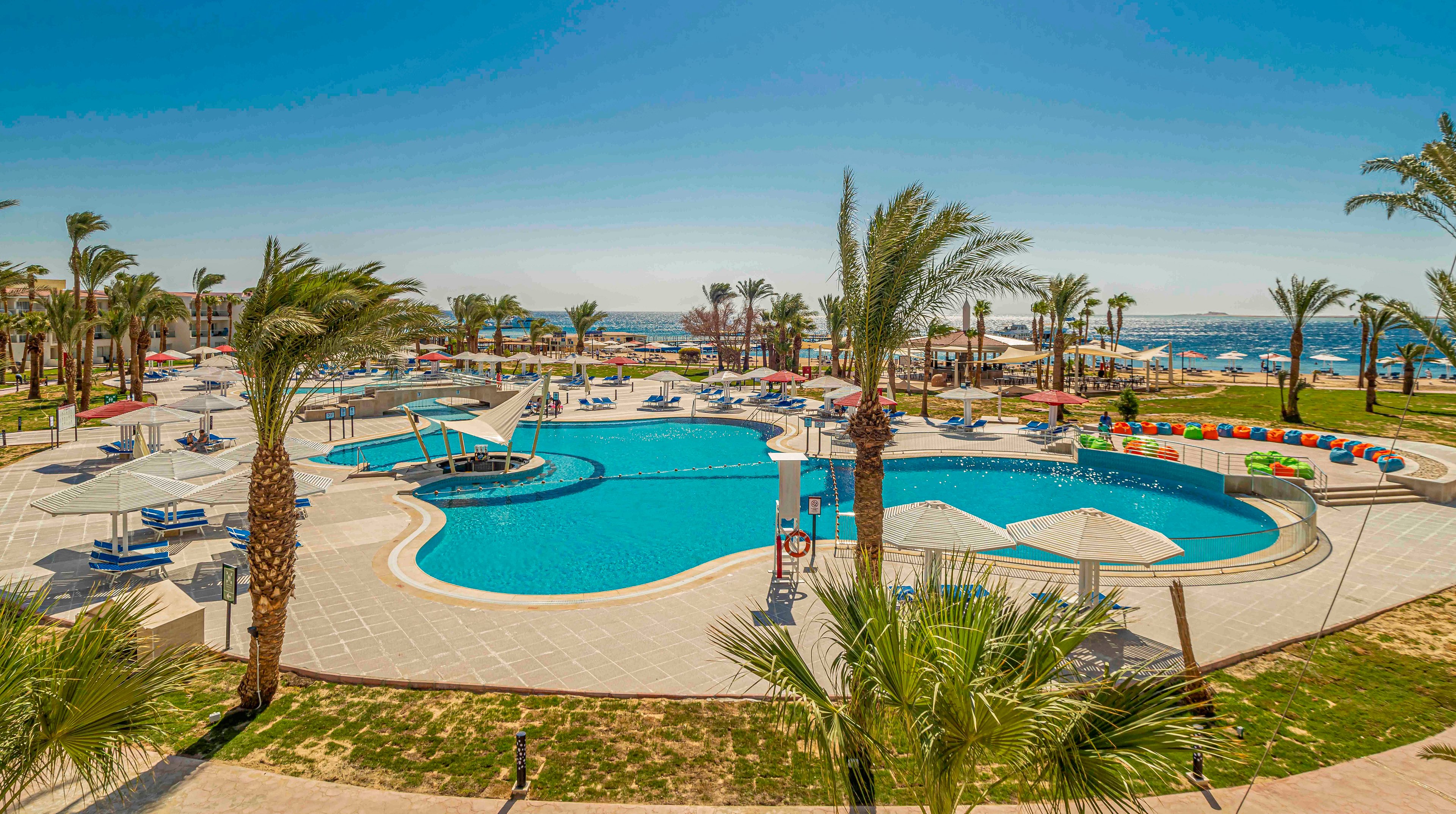 A large resort swimming pool is surrounded by tall palm trees and sun loungers under white umbrellas. The scene overlooks a bright blue sea in the background, suggesting a coastal vacation destination. Colorful seating and shaded areas create a relaxed leisure atmosphere around the pool. The clear sky and strong sunlight emphasize a warm, tropical climate ideal for holiday travel.