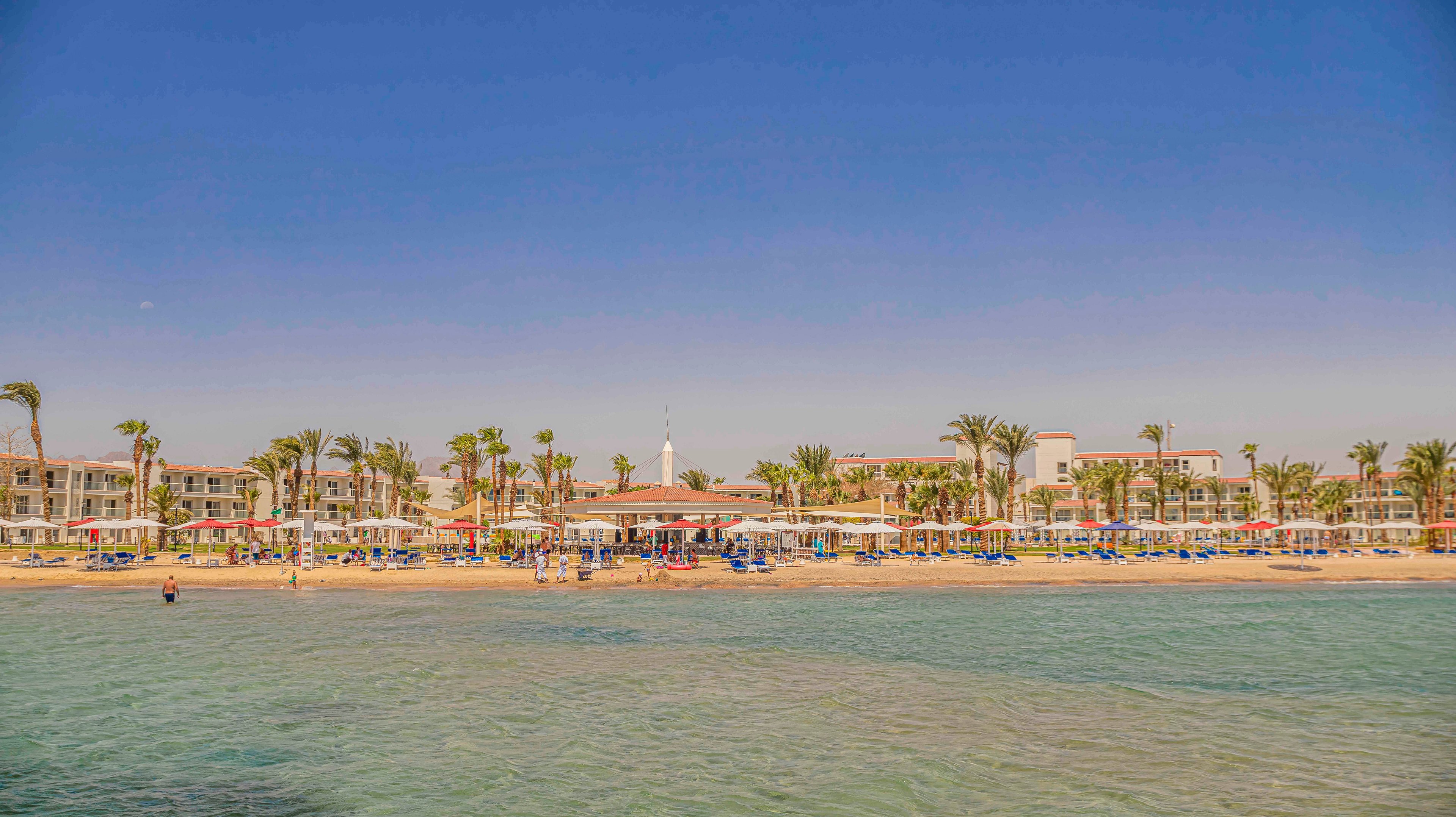 A large beachfront resort stretches along a sandy shore lined with palm trees. Rows of colorful beach umbrellas and sun loungers face the calm, shallow sea. People relax near the water and on the beach under a clear blue sky. The scene suggests a warm, sunny vacation destination with tranquil coastal views.