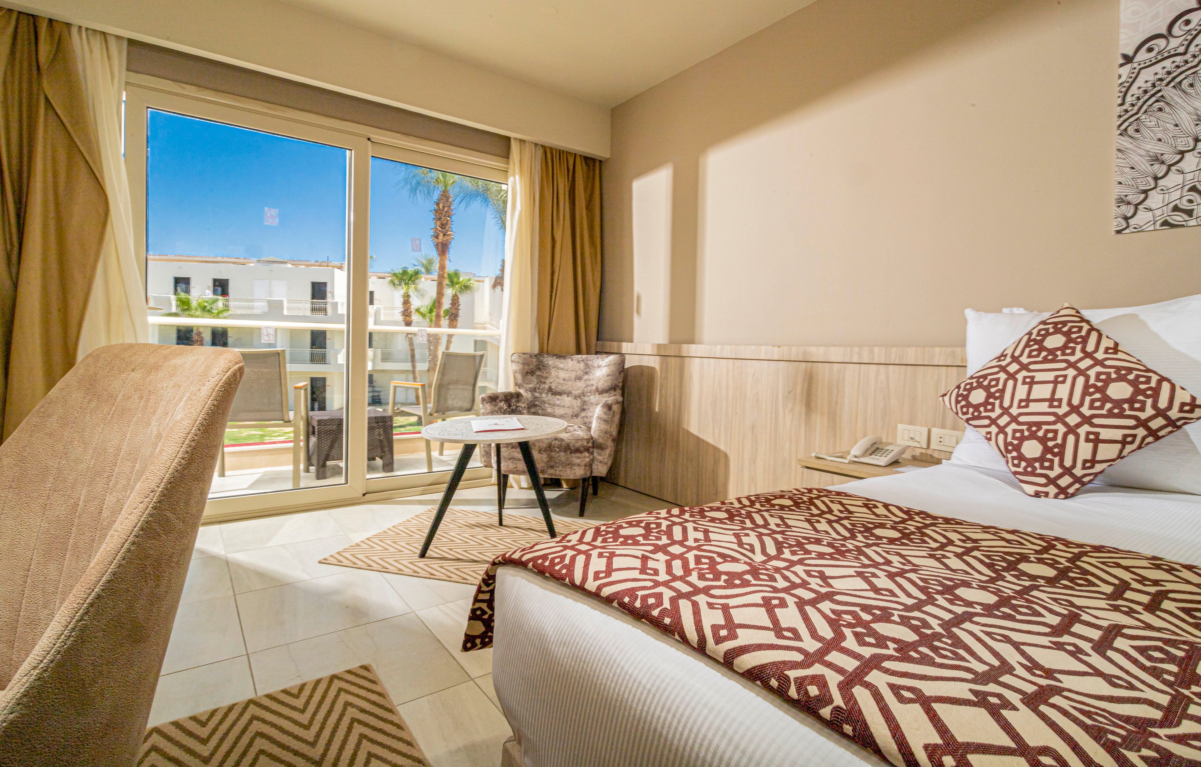 Bright hotel room interior featuring a neatly made bed with patterned linens and cushions. A small seating area with two chairs and a round table is positioned near large sliding glass doors. The doors open to a balcony overlooking a courtyard with palm trees and modern white buildings. Warm neutral tones and natural light create a relaxed, inviting atmosphere suitable for vacation or business travel.