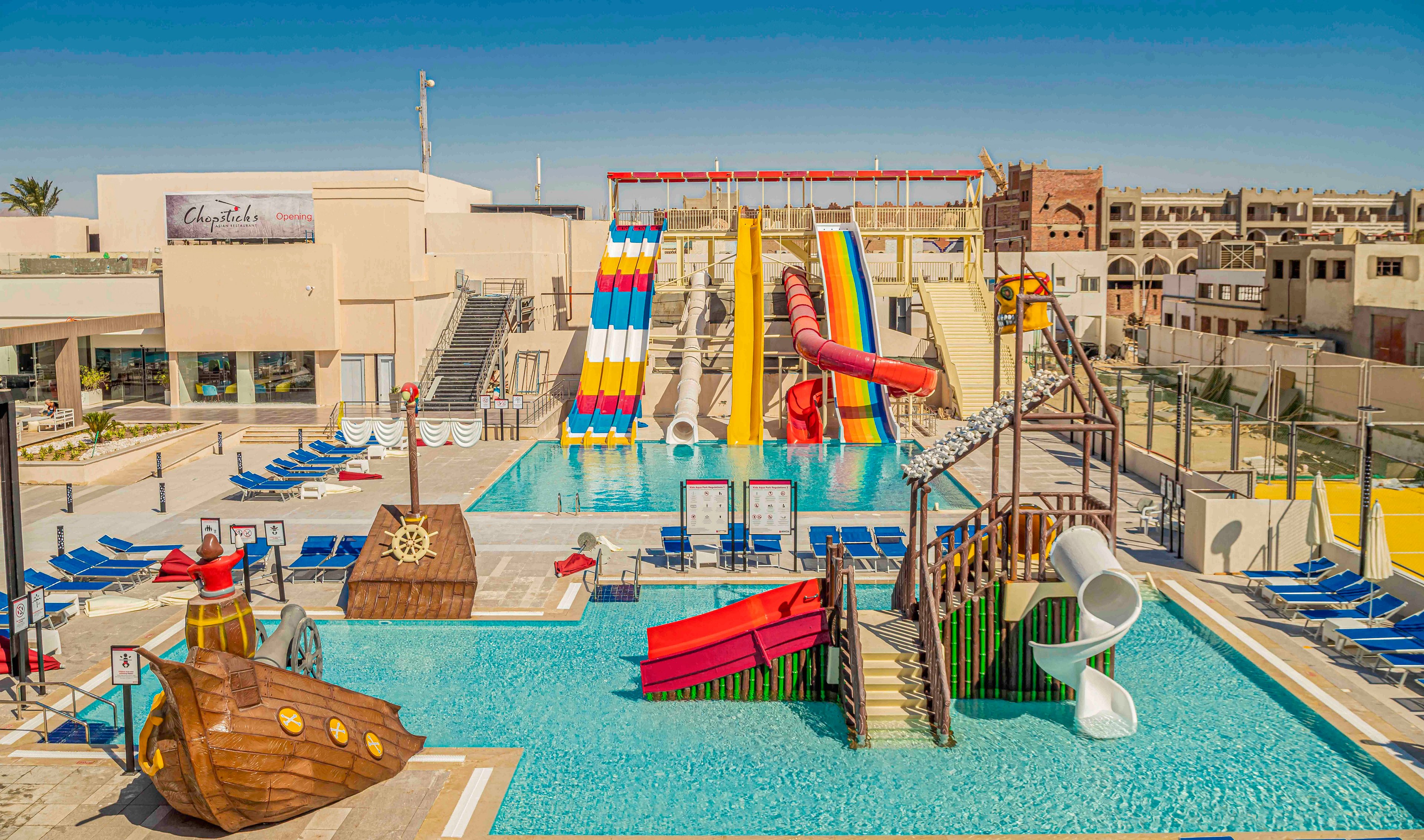 A vibrant resort water park features multiple tall, colorful slides emptying into a large swimming pool. In the foreground, a shallow kids’ pool includes a pirate ship play structure, small slides, and climbing elements. Rows of blue lounge chairs line the pool deck under clear, sunny skies. Surrounding buildings and the main resort complex frame the aquatic area, emphasizing a family-friendly vacation setting.